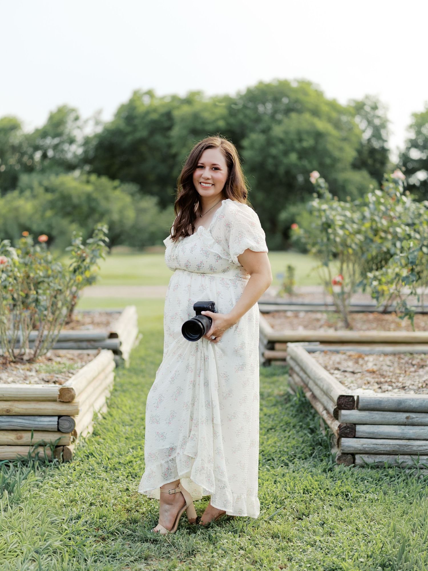 A smiling woman in a white floral dress holds a camera while standing in a lush garden with raised wooden flower beds.