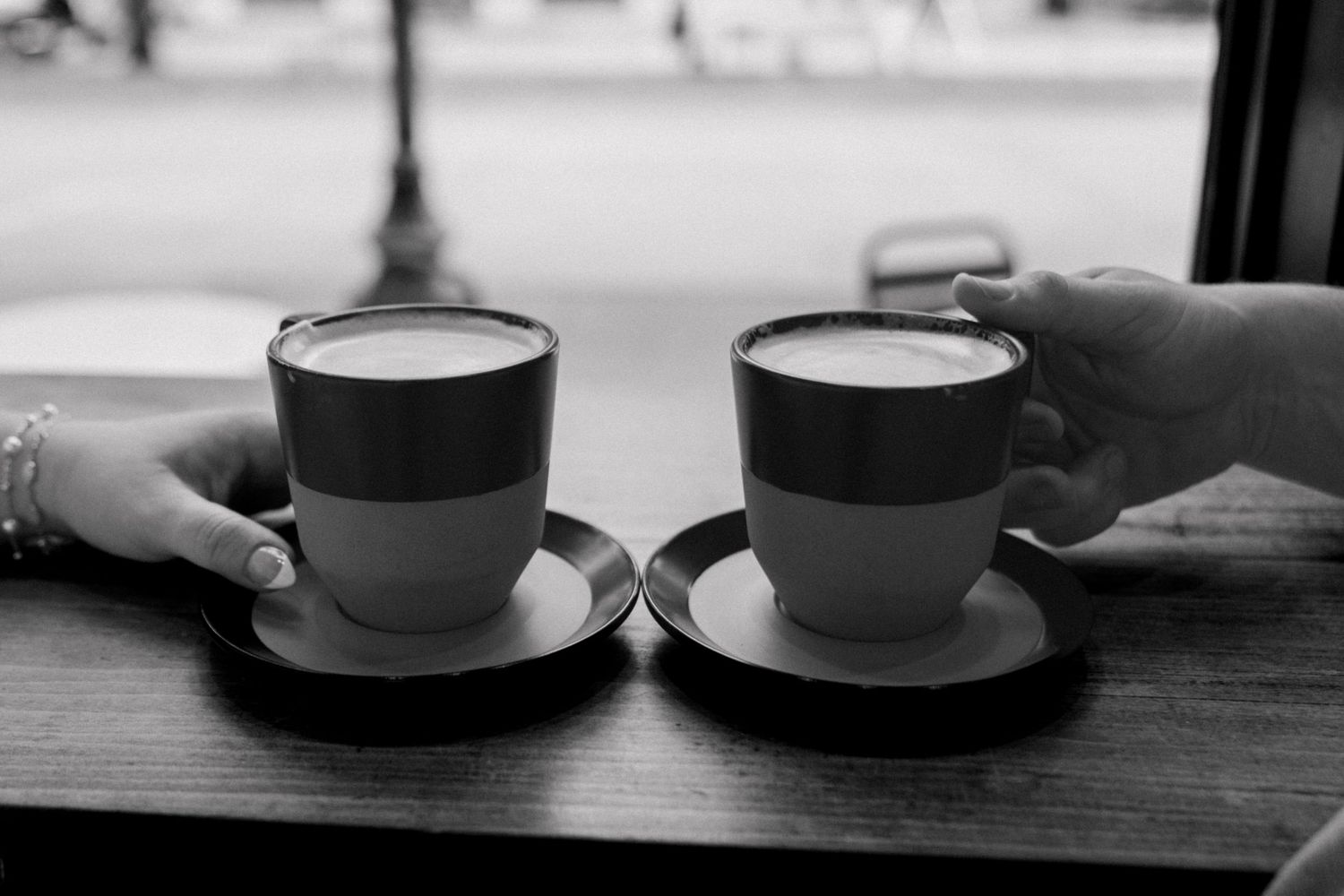 Black and white close-up of two cappuccinos on saucers held by a couple at a cafe table.
