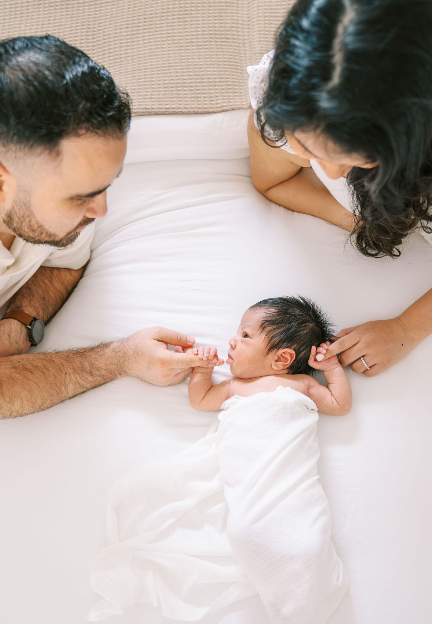 New parents lovingly gaze at their swaddled newborn baby lying on a white bed.