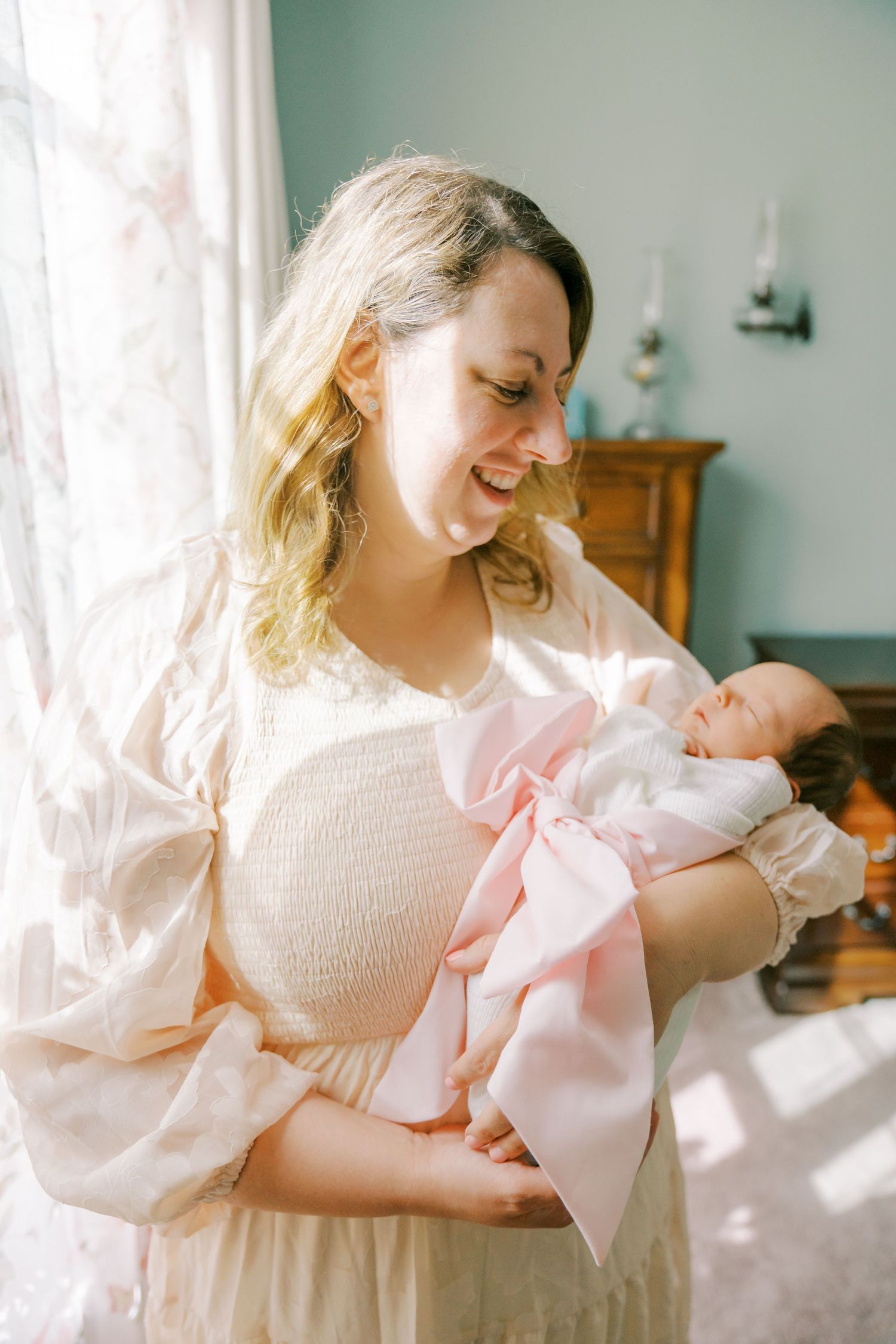 A tender moment in natural light as a woman in cream dress holds a newborn wrapped in pink and white.