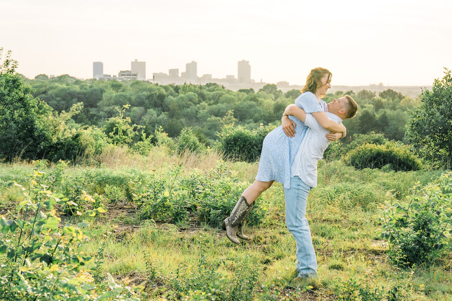 A couple embraces in a meadow with a city skyline visible in the background during golden hour.