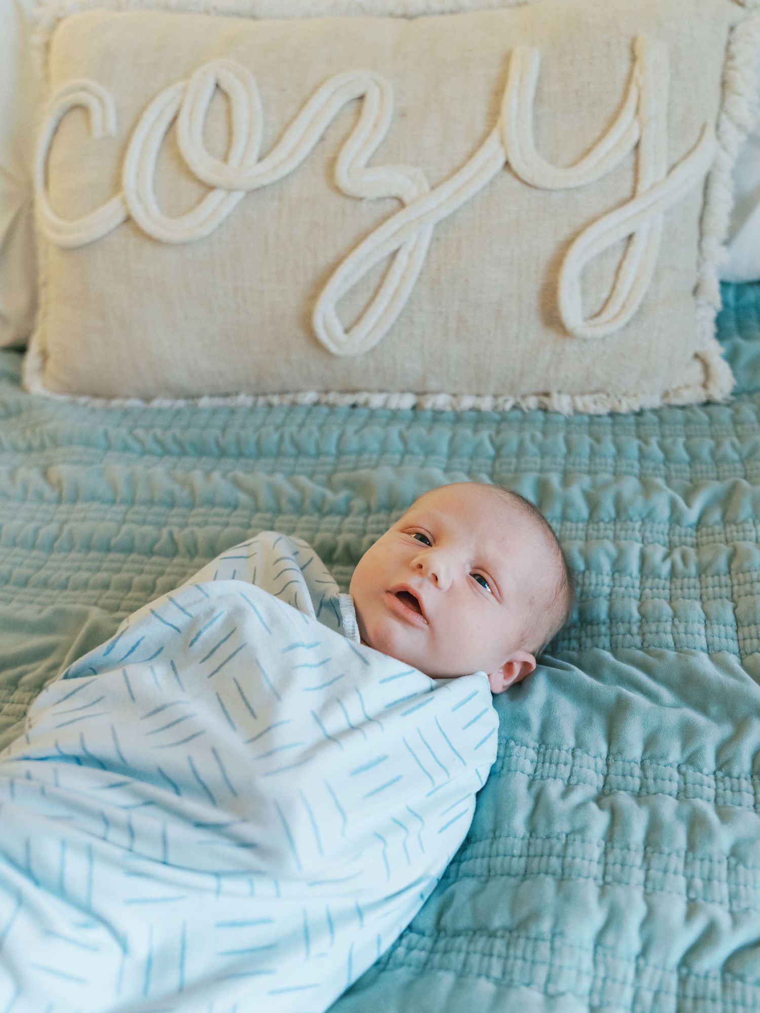 Newborn baby swaddled in white lying on a turquoise quilted blanket with decorative text on wall above.