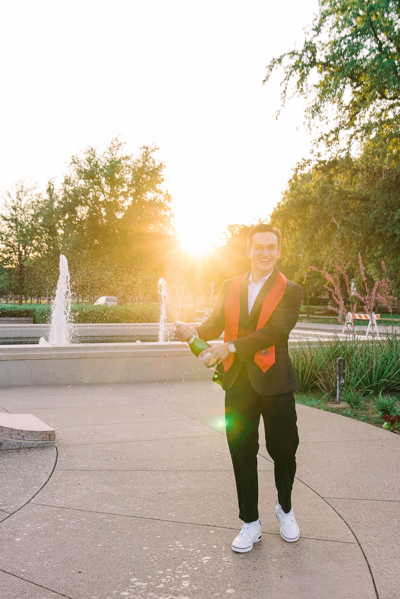 A person in a black suit and white sneakers poses in front of a fountain at sunset creating a warm backlit glow.