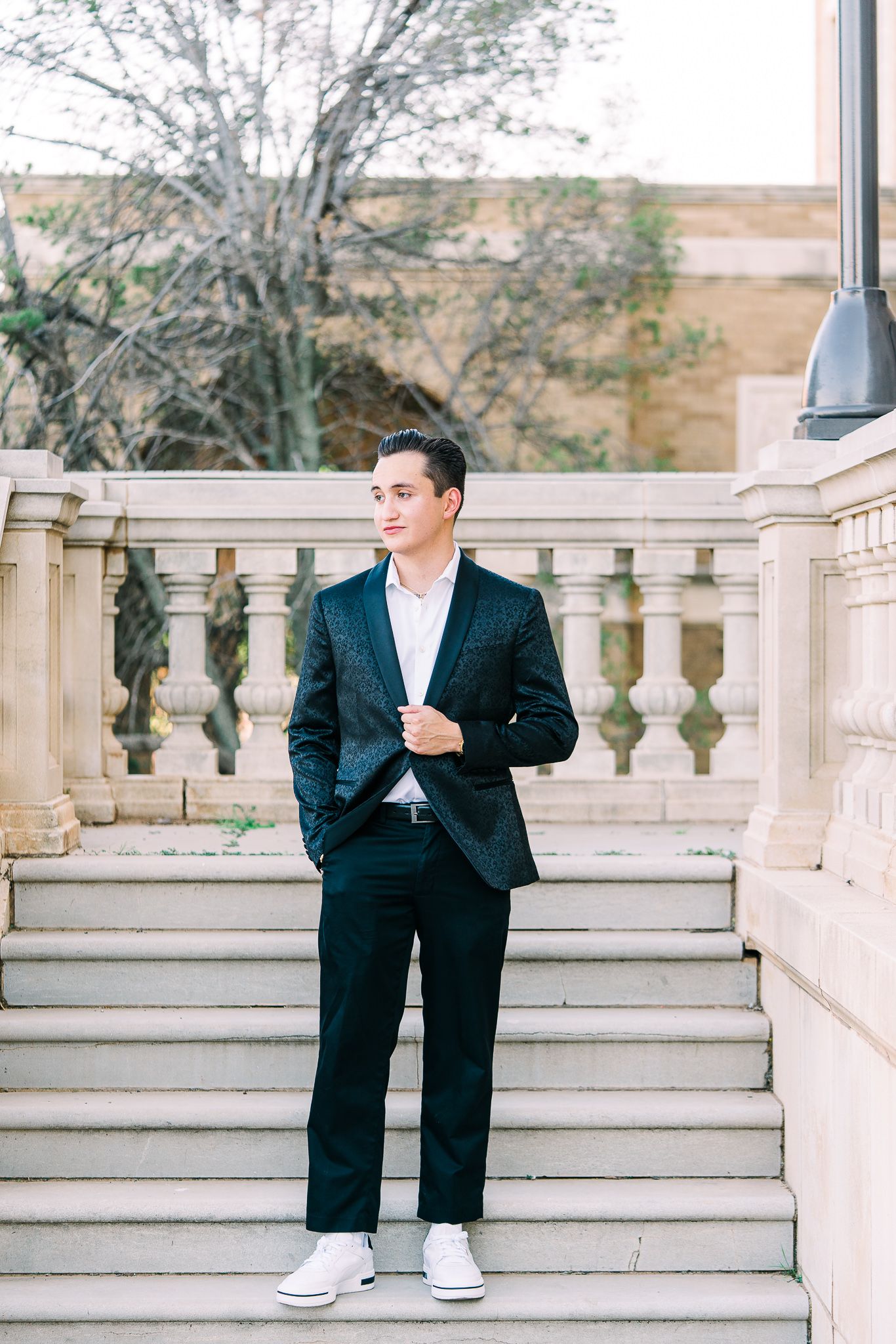A person in formal attire and casual shoes poses on elegant stone steps with decorative railings.
