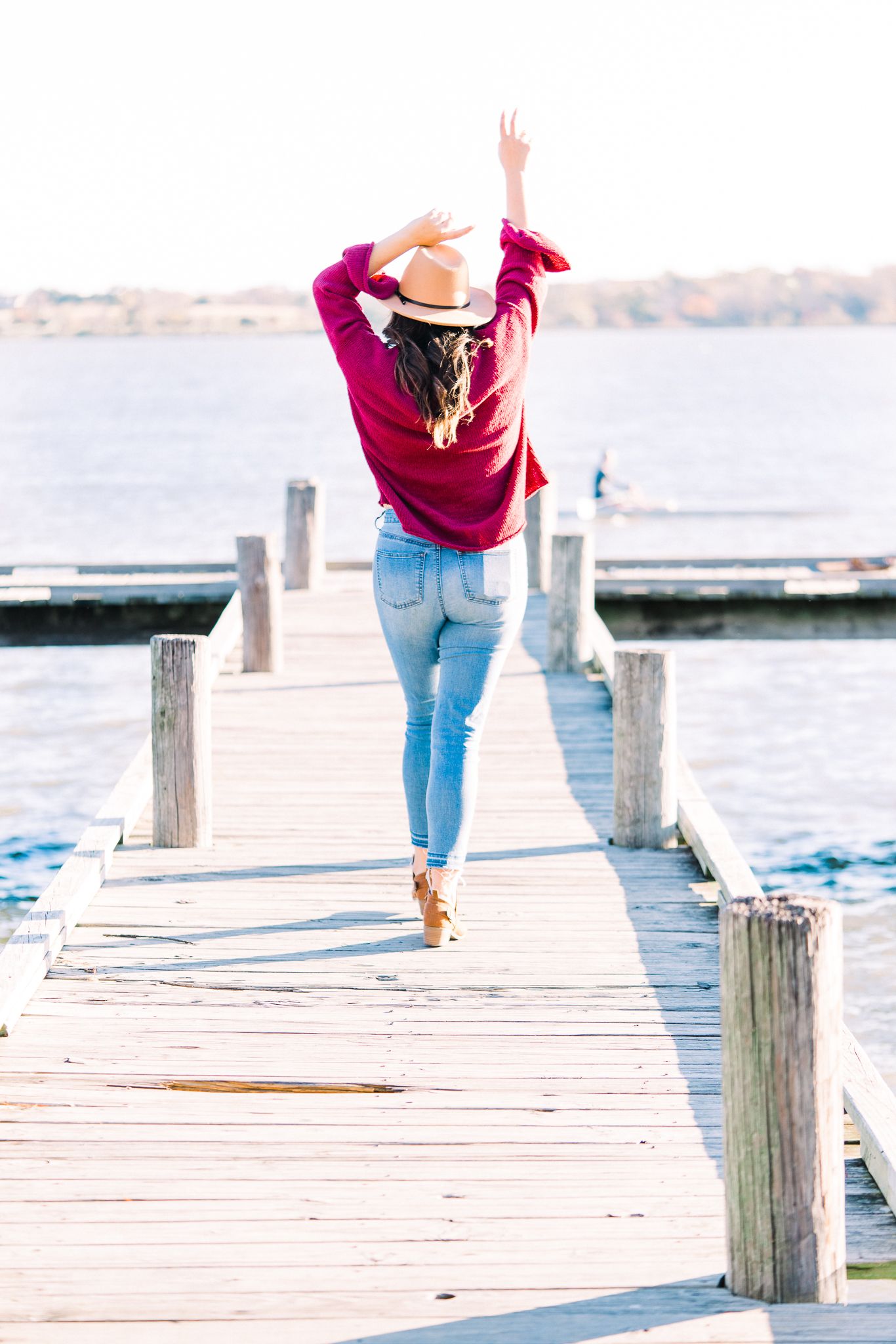 Person in burgundy sweater and jeans walks along wooden dock by waterfront during sunset.