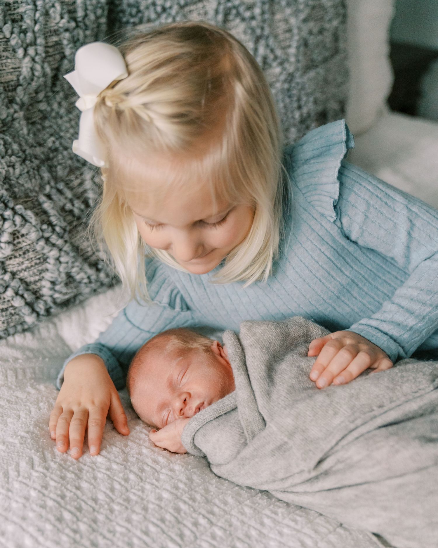 Siblings share a tender moment with newborn baby on grey blanket.