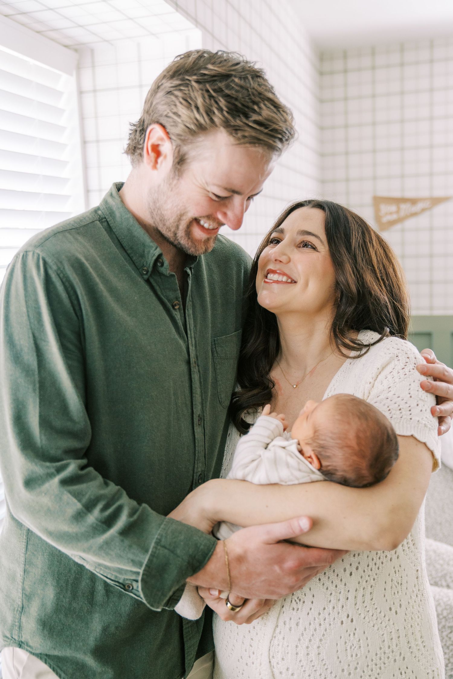 A couple smiles lovingly while holding their newborn baby in a bright room with white decor.