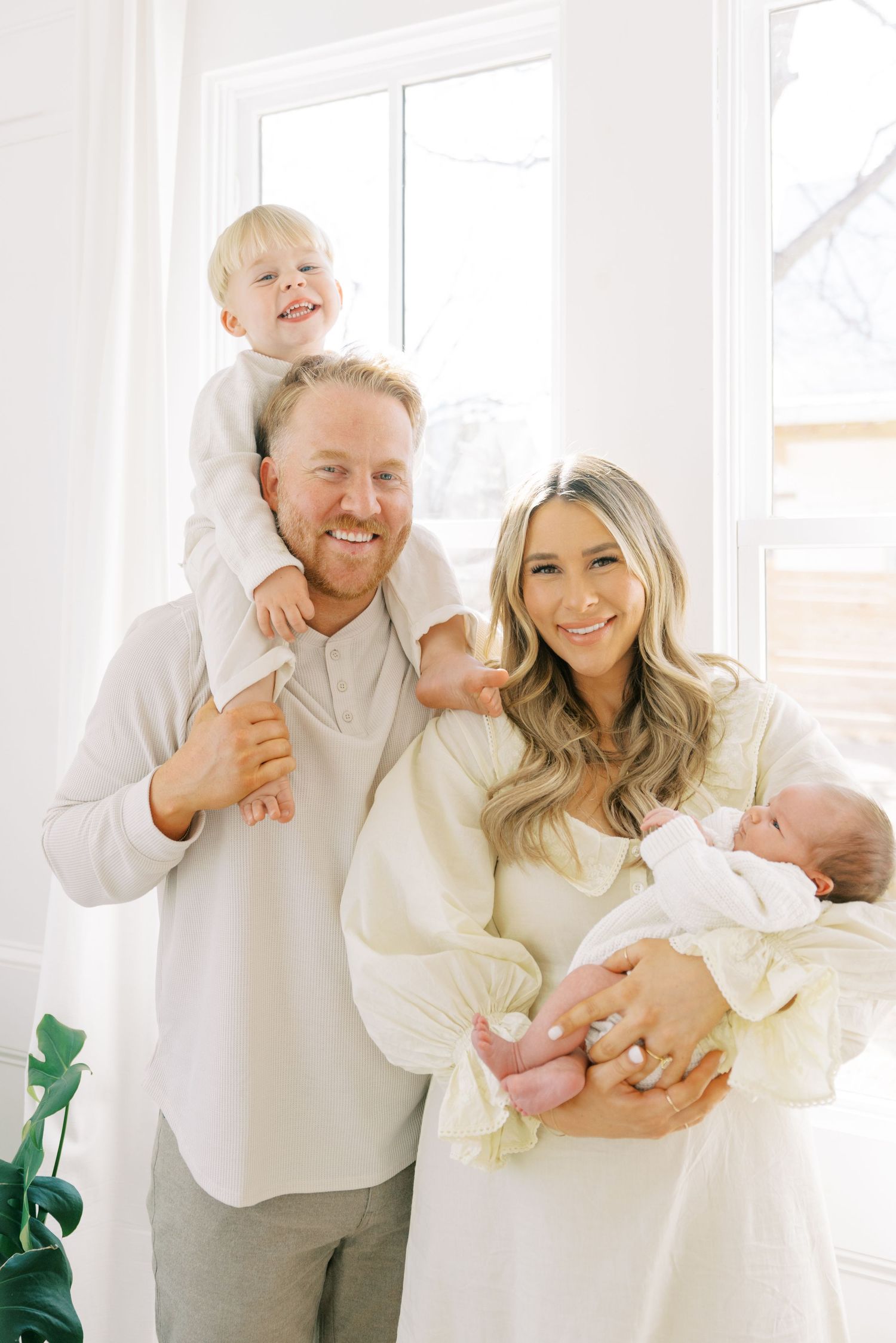 Happy family of four posing indoors, father holding toddler on shoulders, mother cradling newborn baby, wearing white outfits.