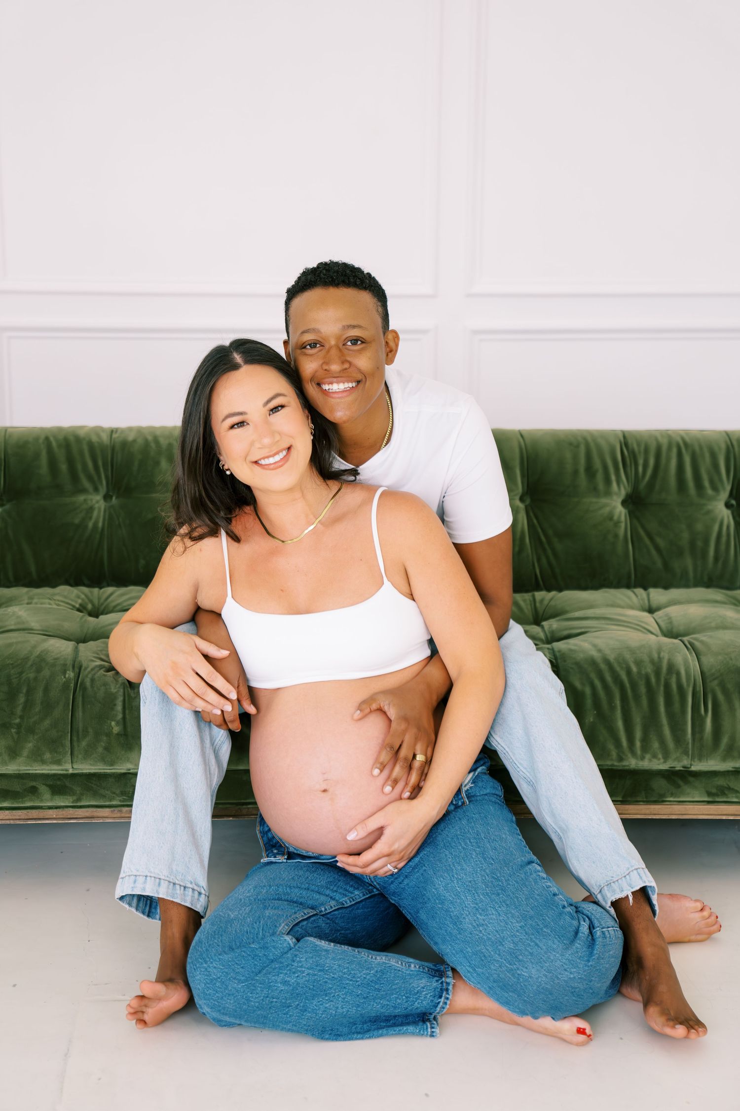A happy interracial couple poses for a maternity photo, sitting against a green velvet sofa, cradling the pregnant woman's bare belly.
