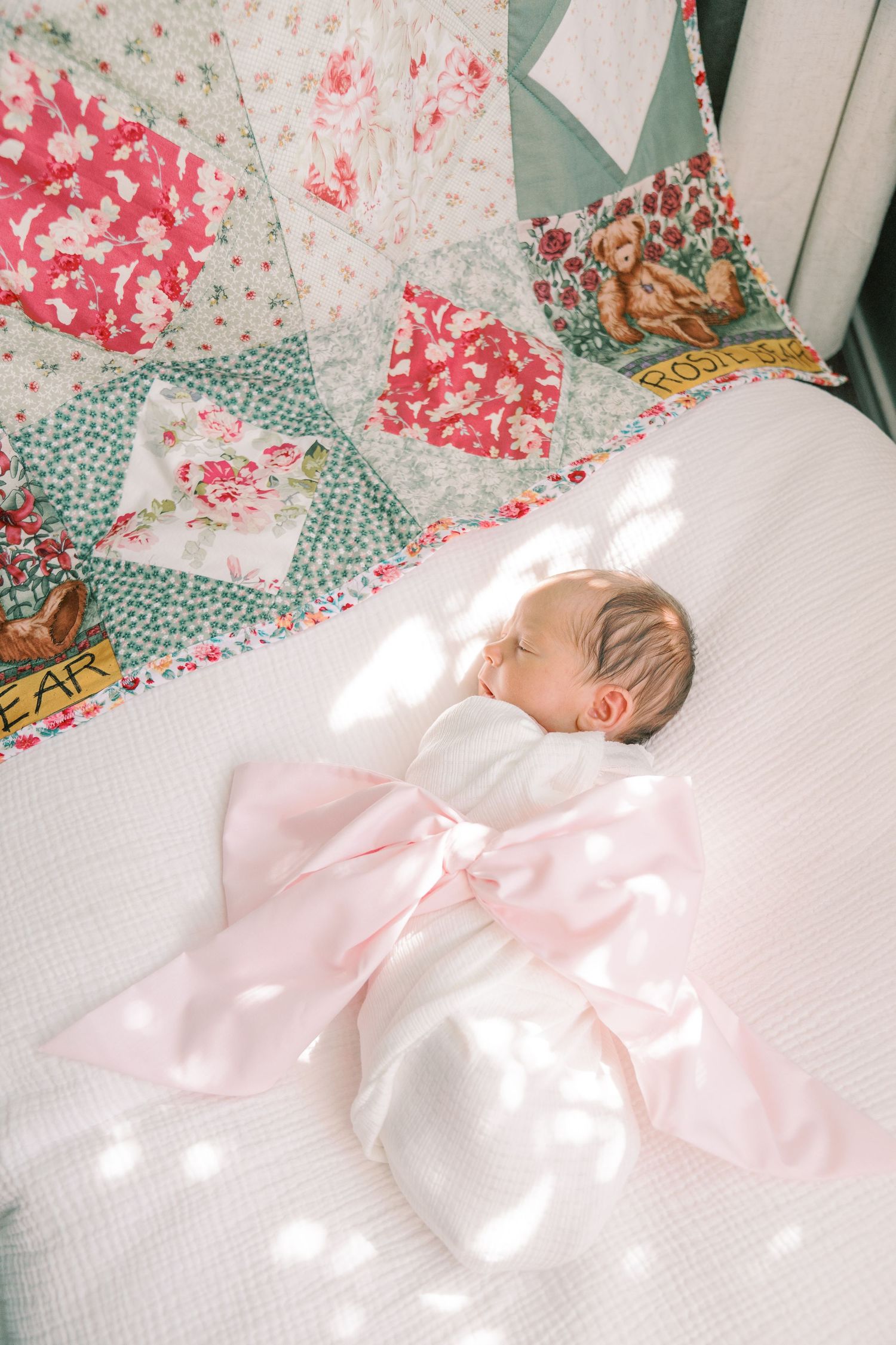 A sleeping newborn baby wrapped in white with a pink bow lies on a colorful patchwork quilt in sunlit room.