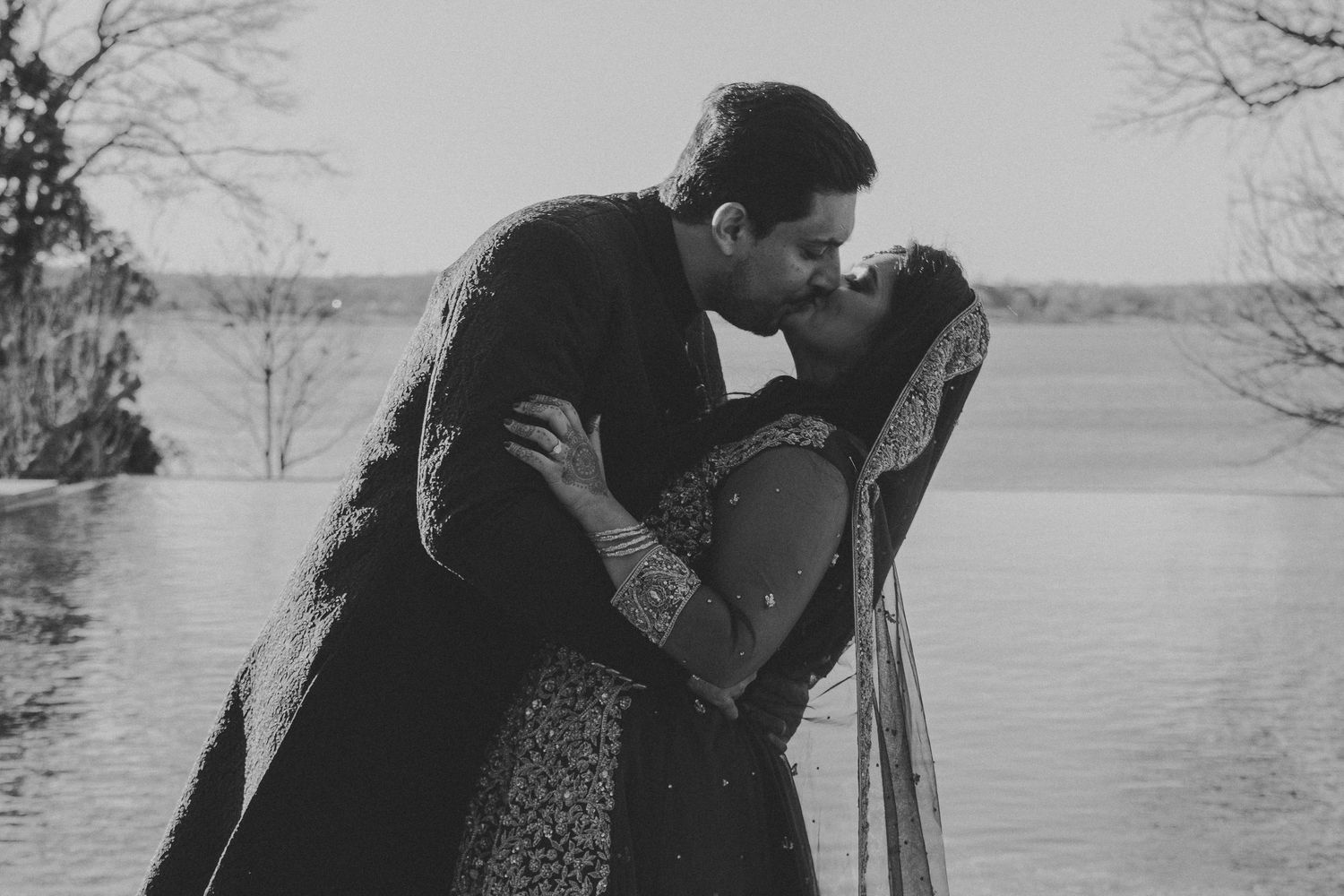 Black and white photo of couple embracing by a lake with bare trees in the background.