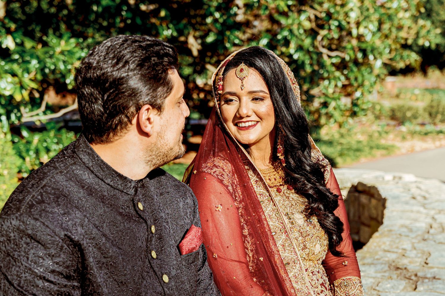 A couple in traditional red Indian wedding attire share intimate moments during an outdoor photoshoot.