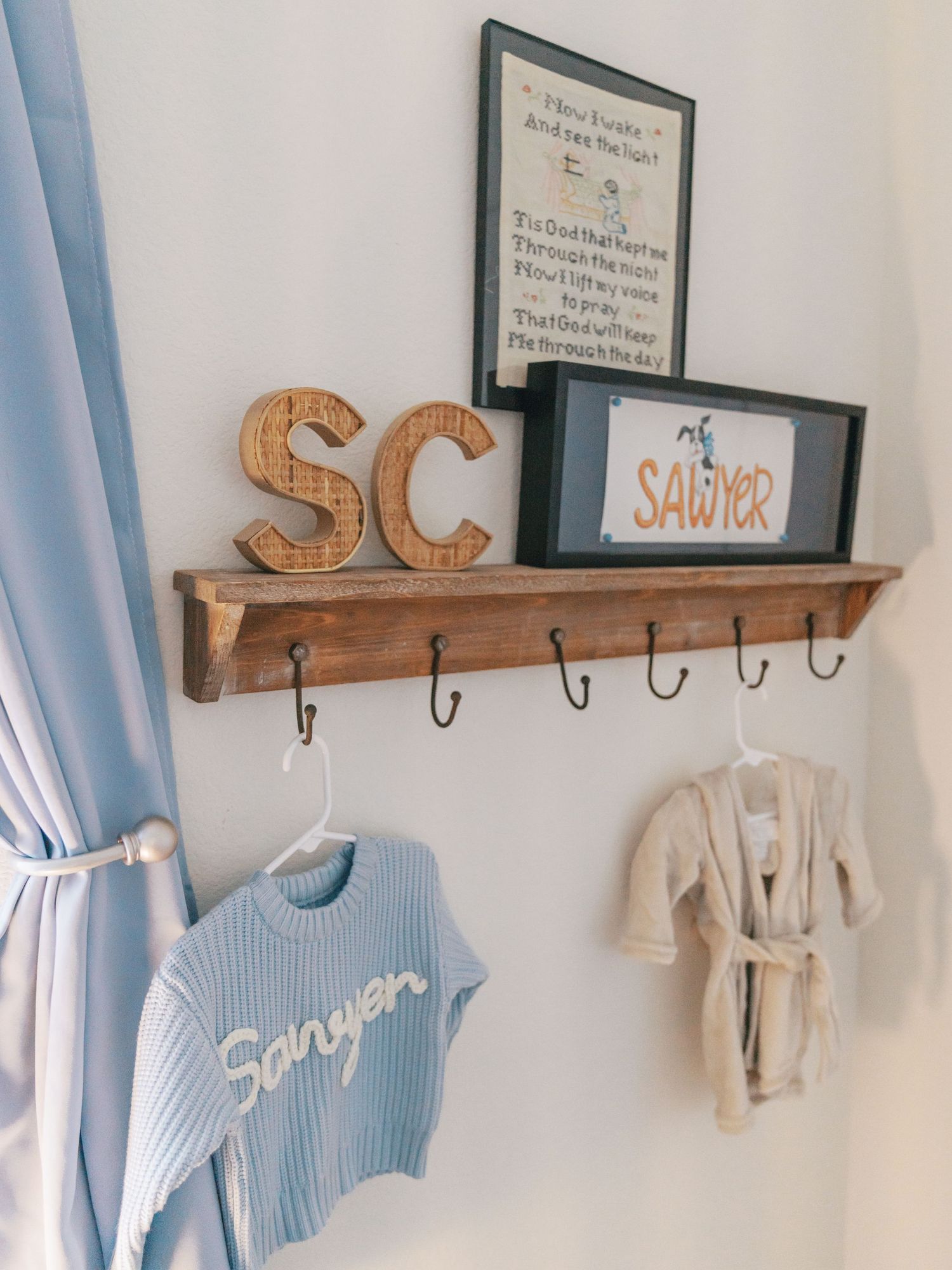 A wooden coat rack with hooks displays baby clothes and nursery decor against a white wall with blue curtains.