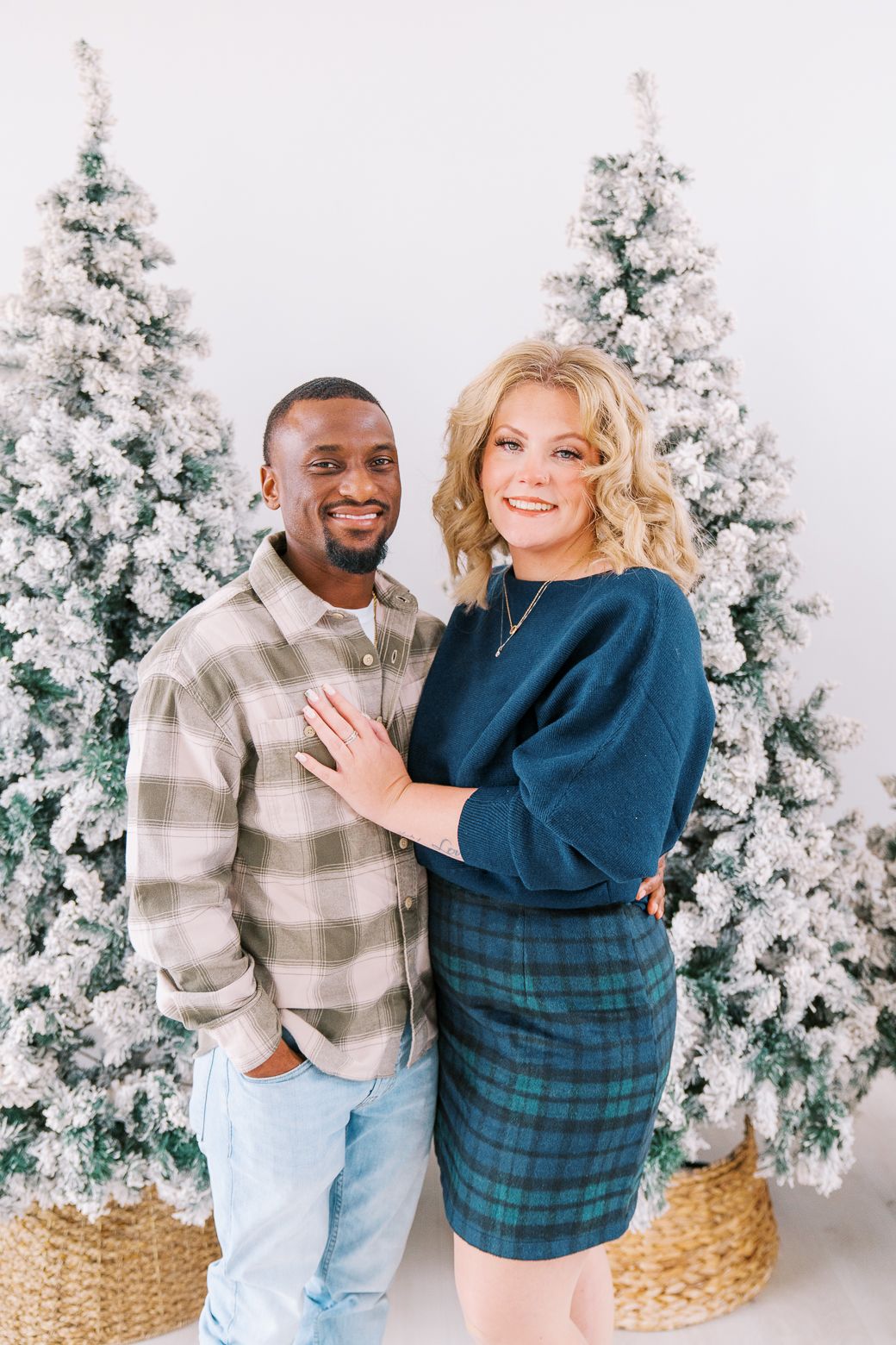 A couple poses together in front of snow-covered Christmas trees in a festive holiday photoshoot.