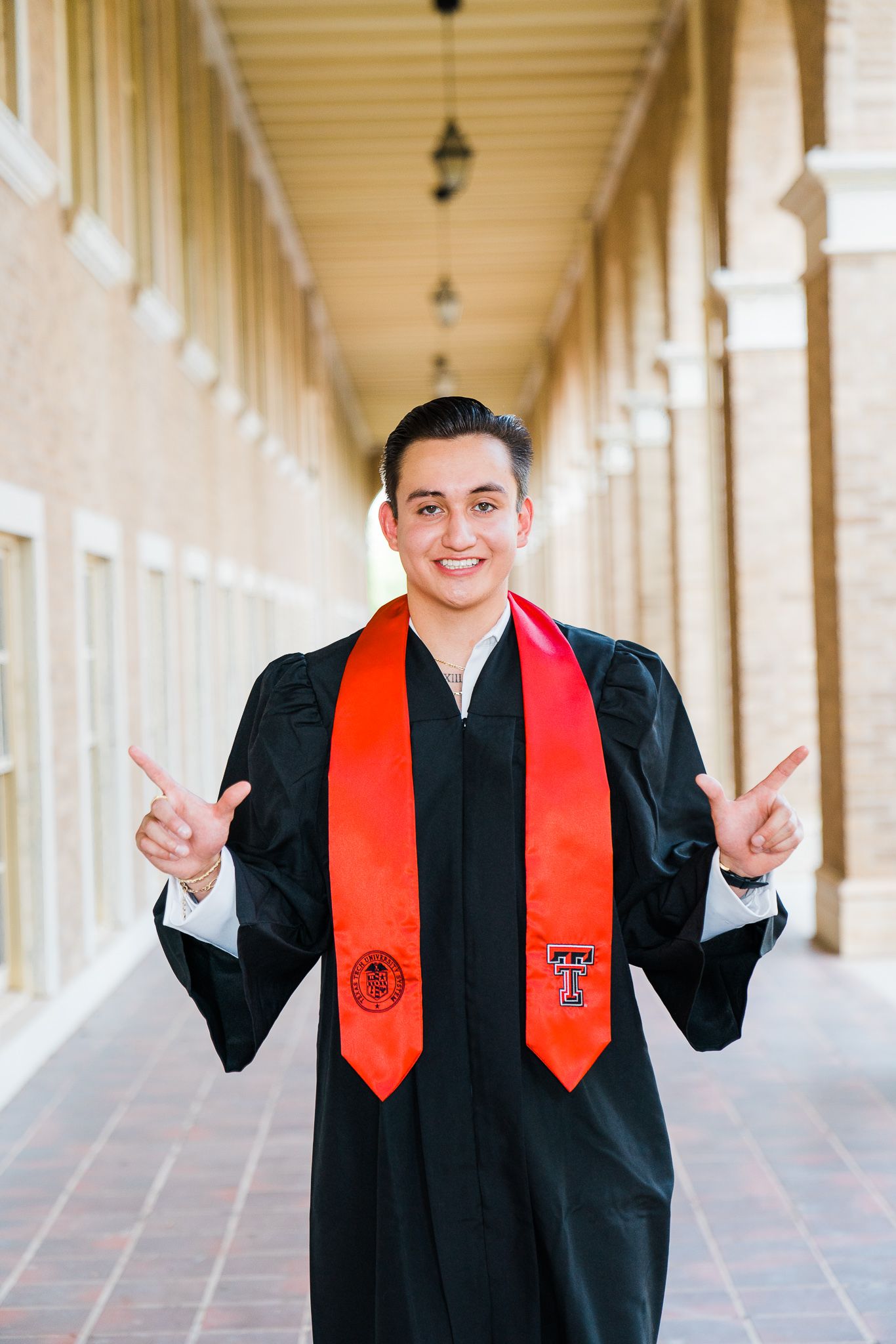 Graduate in black cap and gown with red stole making peace signs in a Spanish colonial style hallway.