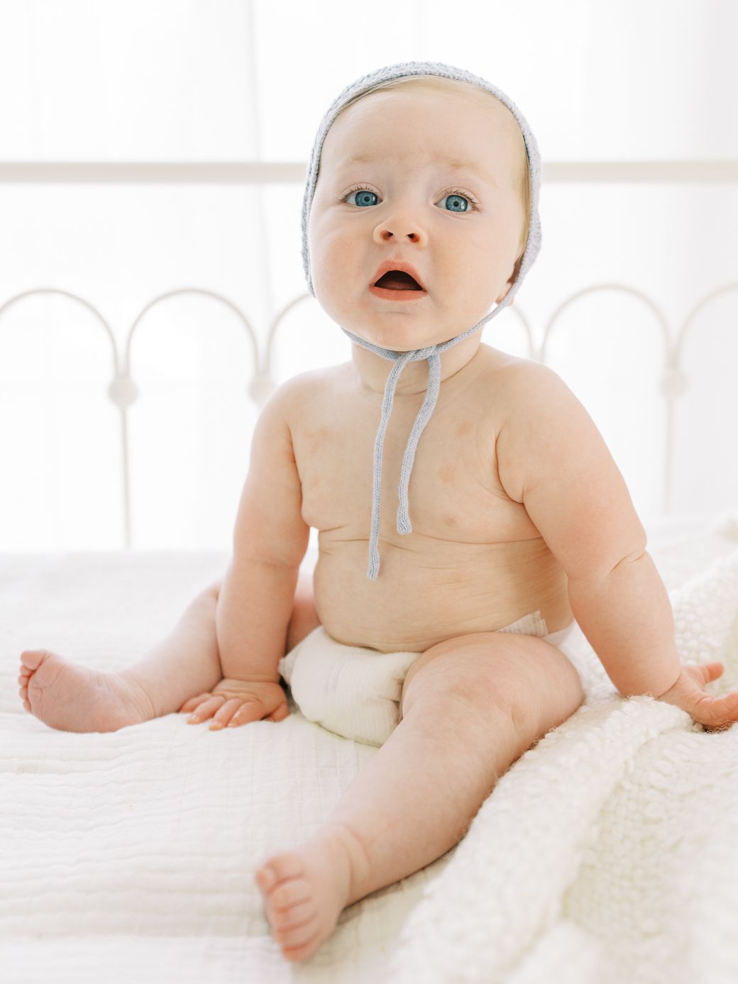 Baby wearing a light colored bib lies on white bedding in a bright sunny room.