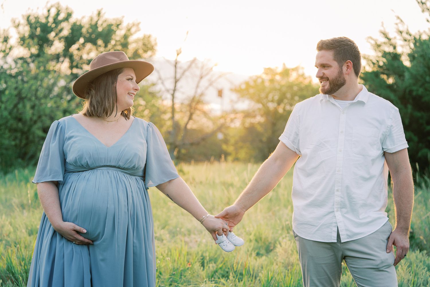 Expecting couple walks hand in hand through sunlit meadow at golden hour.