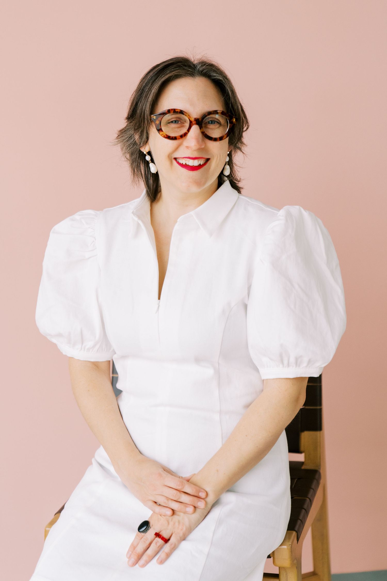 A person in a white puff sleeve top and red glasses poses against a pink background in a studio setting.