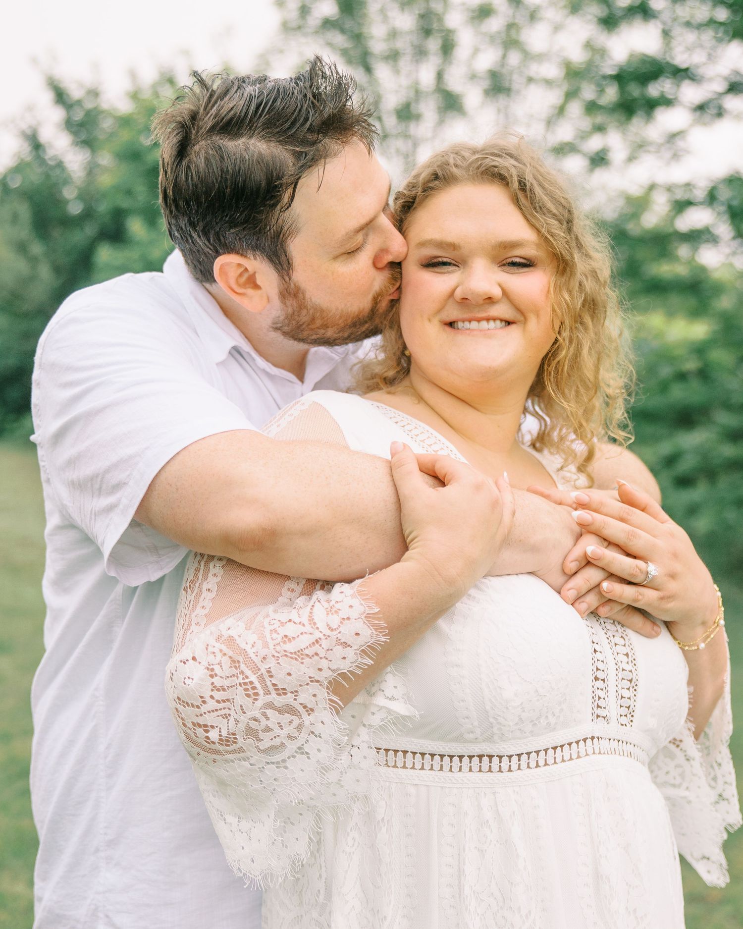 Man in white shirt kissing woman on cheek while embracing her from behind outdoors in garden setting.
