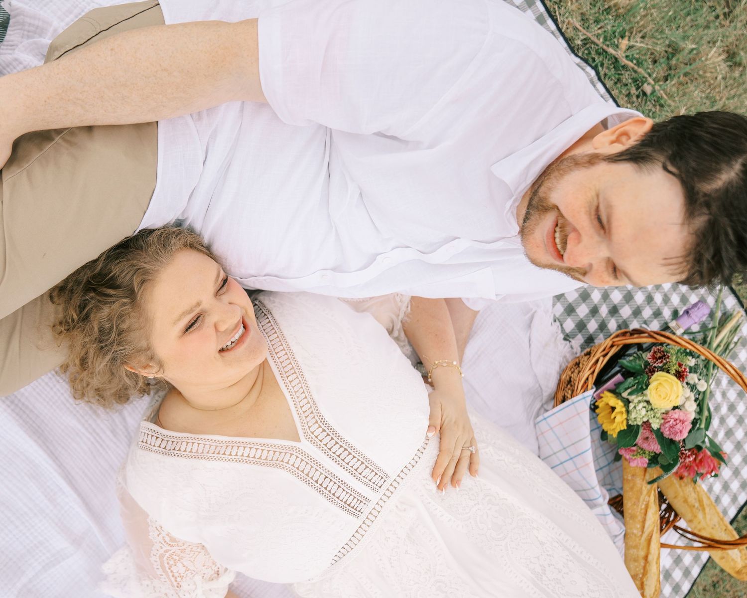 Happy couple lying on white blanket outdoors laughing together near a picnic basket with flowers.