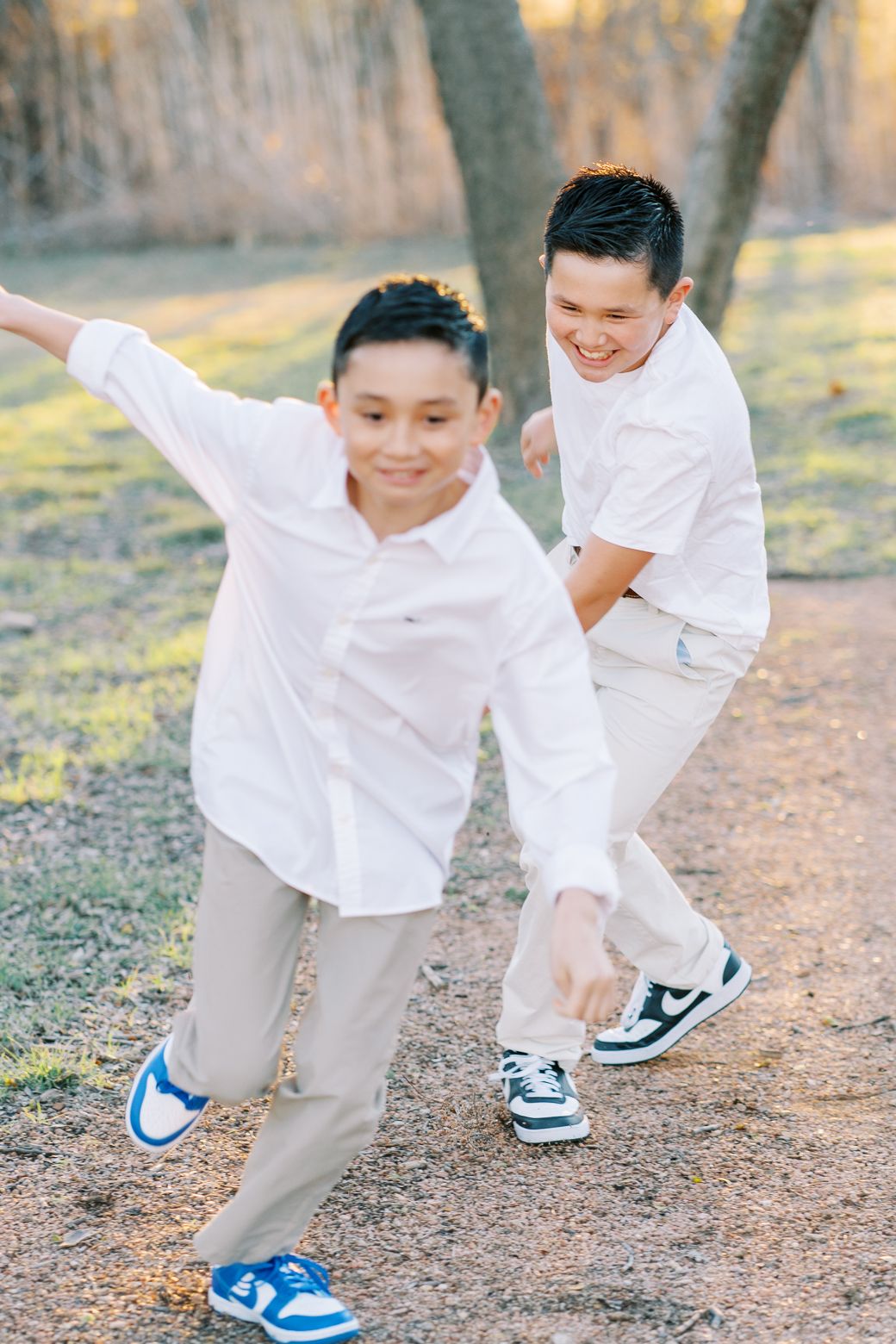 Two boys in white shirts and khaki pants play outdoors, smiling and posing energetically on a dirt path.