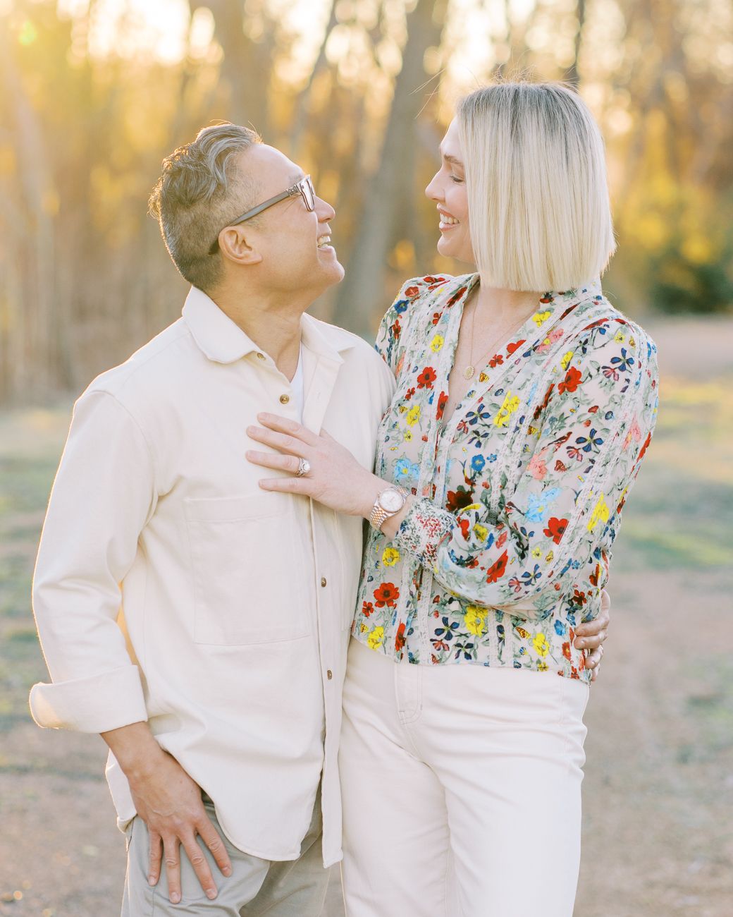 Couple smiling at each other outdoors at golden hour, woman in floral blouse and man in white shirt.