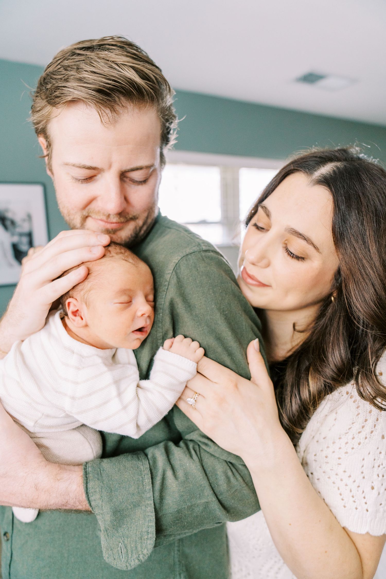 A tender family moment as parents share a loving embrace with their sleeping newborn baby in a bright home setting.