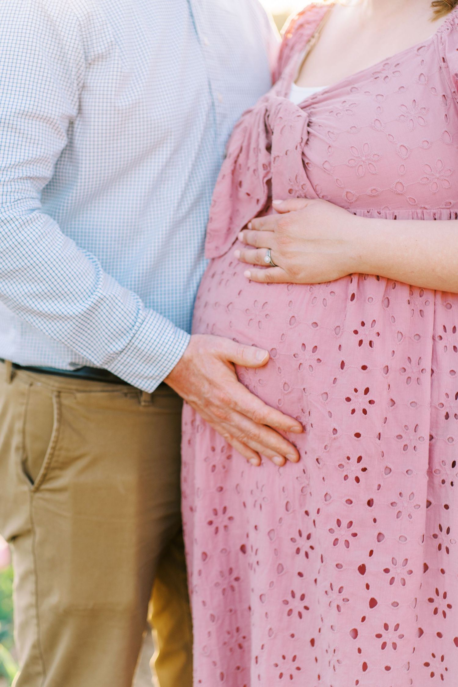Couple in maternity photoshoot, man in blue shirt touching pregnant womans pink dress belly outdoors.