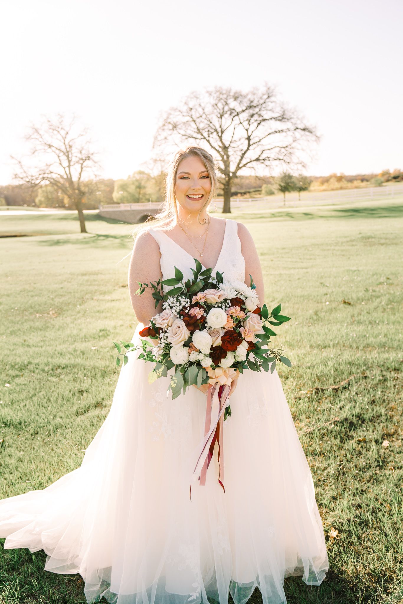 Bride in flowing white tulle gown poses in golden sunlit field with romantic burgundy and blush floral bouquet.