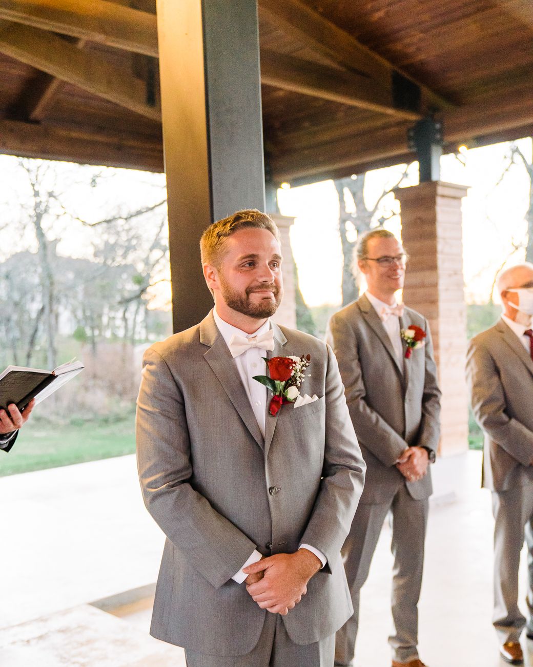 Groomsmen in matching gray suits stand at a covered outdoor venue during a wedding ceremony.