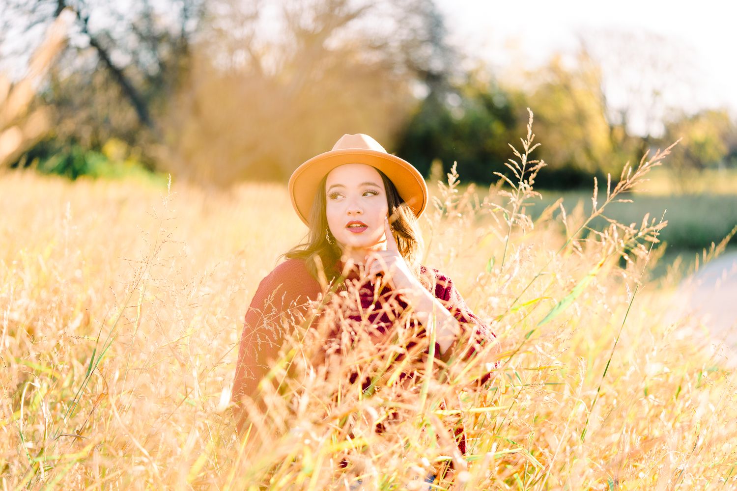 A figure in a wide-brimmed hat sits among golden autumn grass during a sunny afternoon photoshoot.