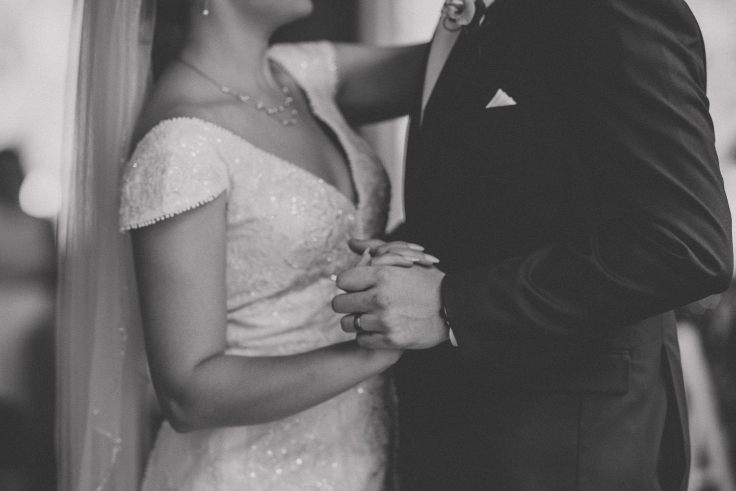 A black and white wedding photo showing a close-up of a couple holding hands, with the bride in a lace dress.
