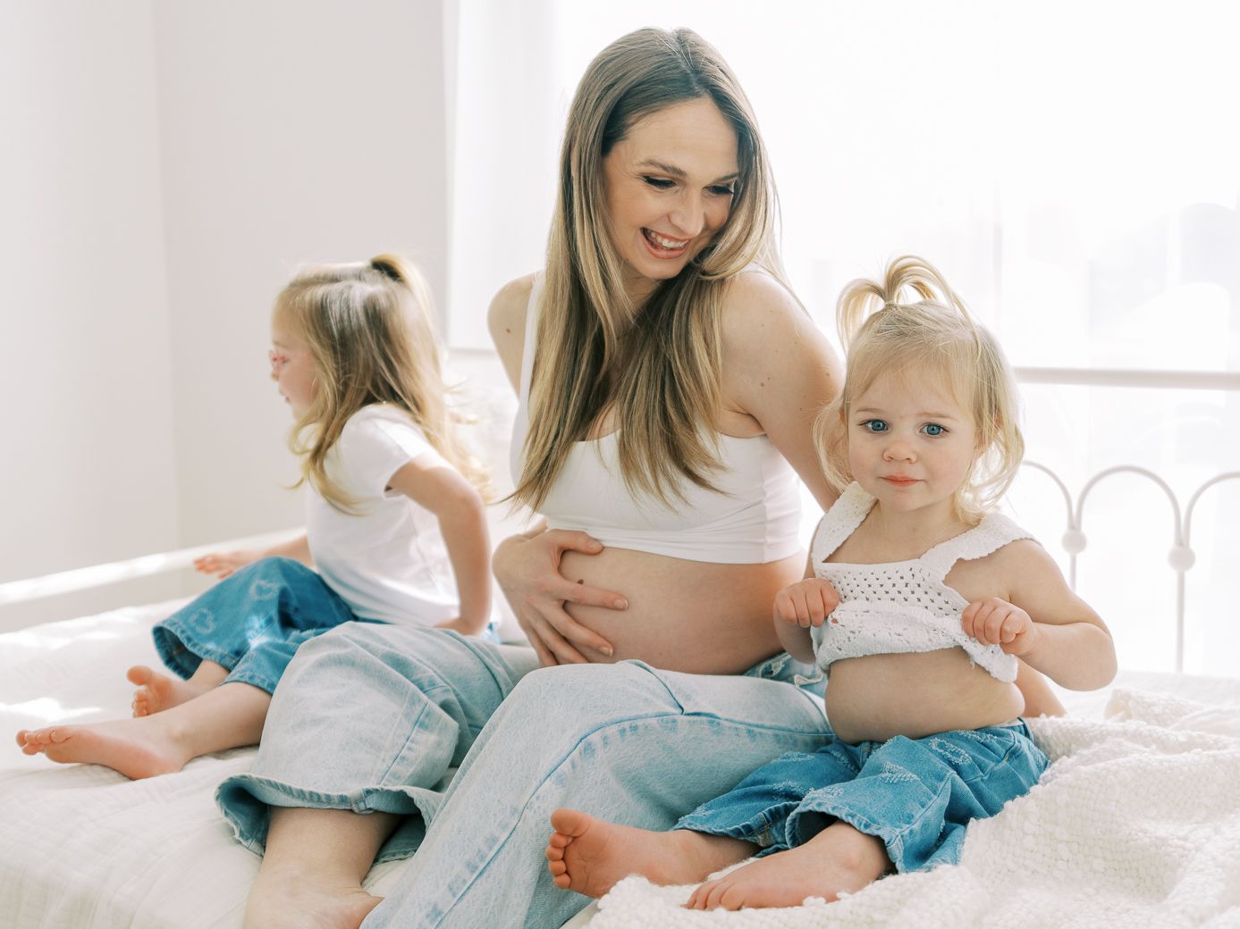 A tender family moment captured in bright white bedroom showing maternal bonding and playful interaction.
