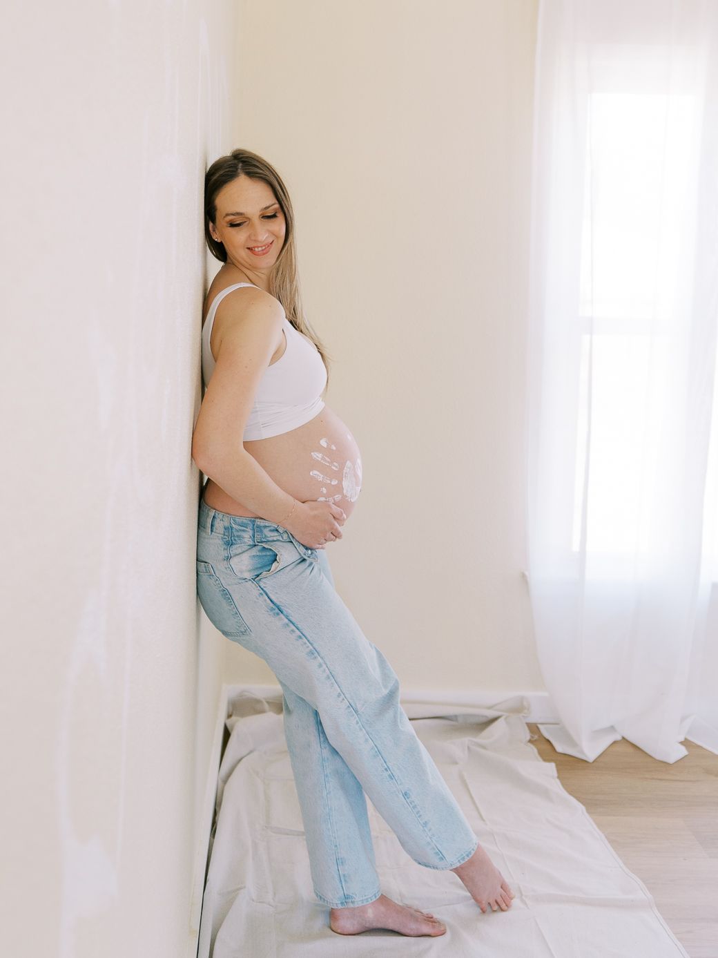 Expectant mother in white top and light jeans poses against bright window.
