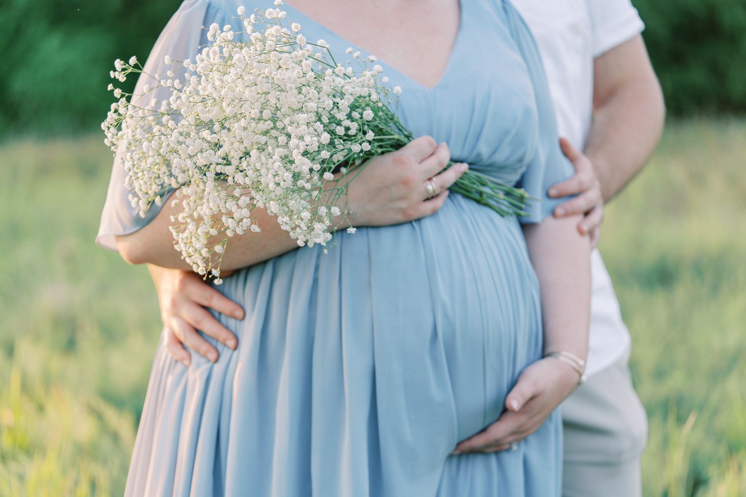 A pregnant figure in a flowing light blue dress holds a delicate bouquet of baby's breath flowers in a sunlit field.