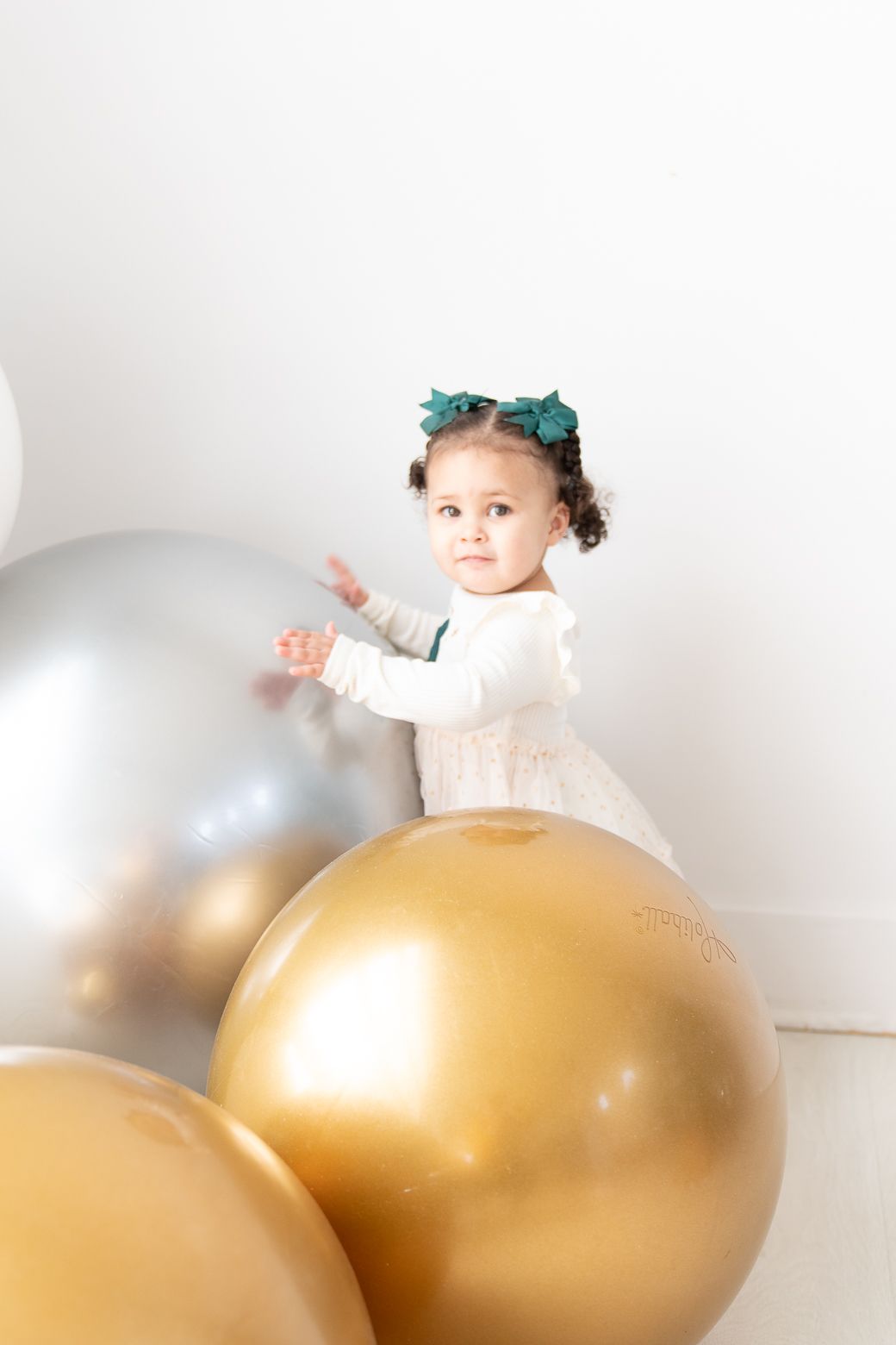 Playful scene with child surrounded by oversized white and gold balloons against white background.