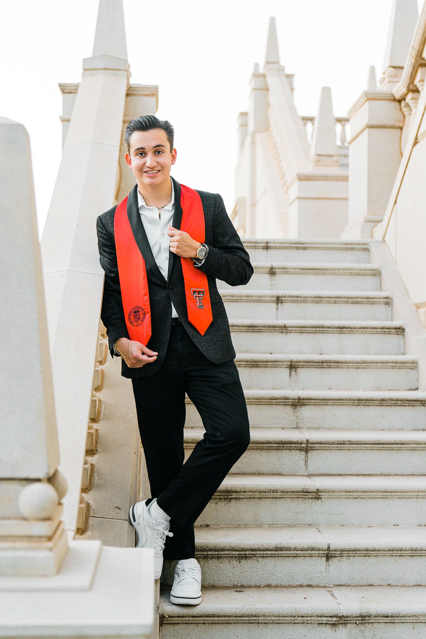 Man in black suit with red stole poses playfully on white stone steps.