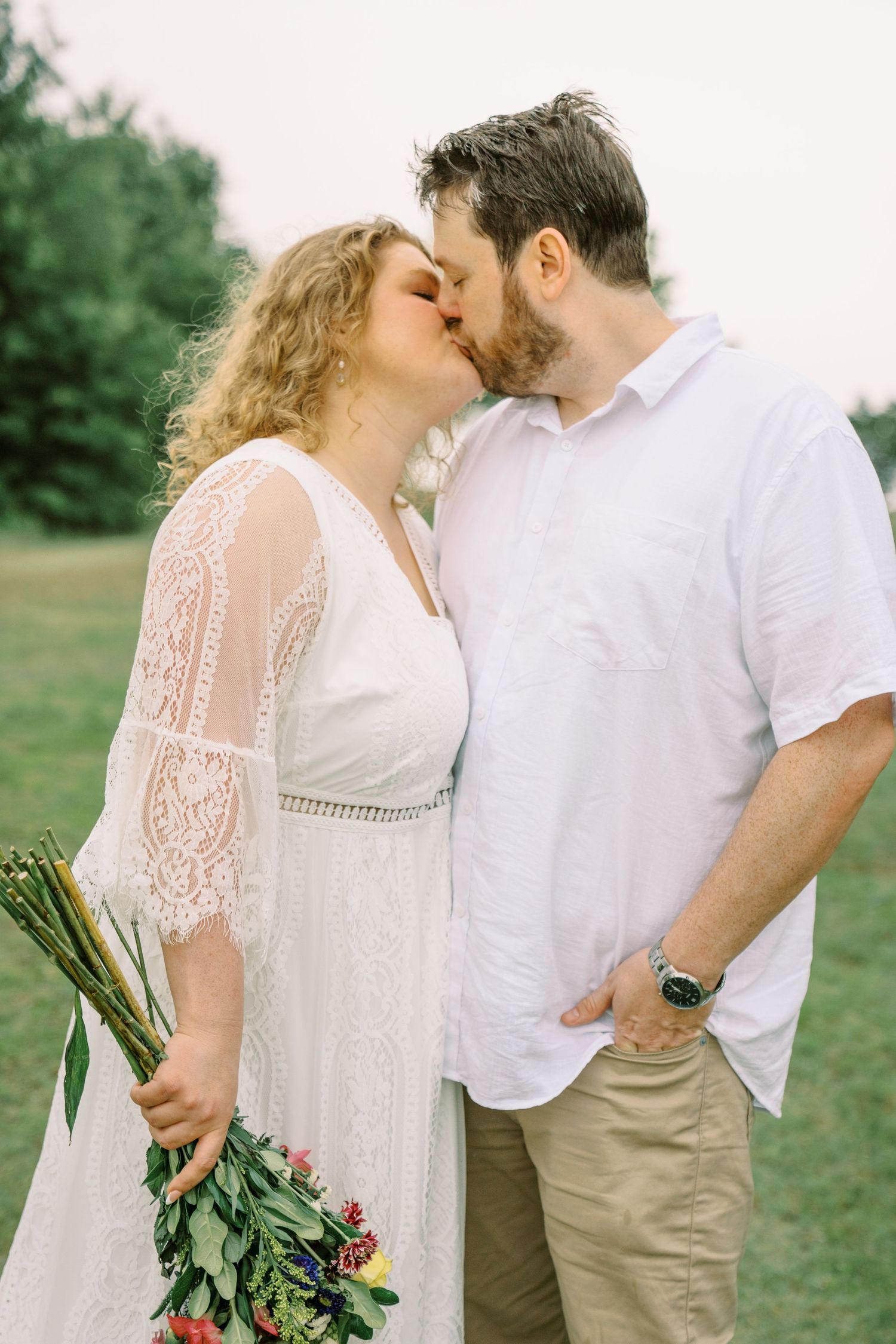A bride in a lace dress and groom in white shirt share a romantic kiss outdoors, as she holds a colorful bouquet.