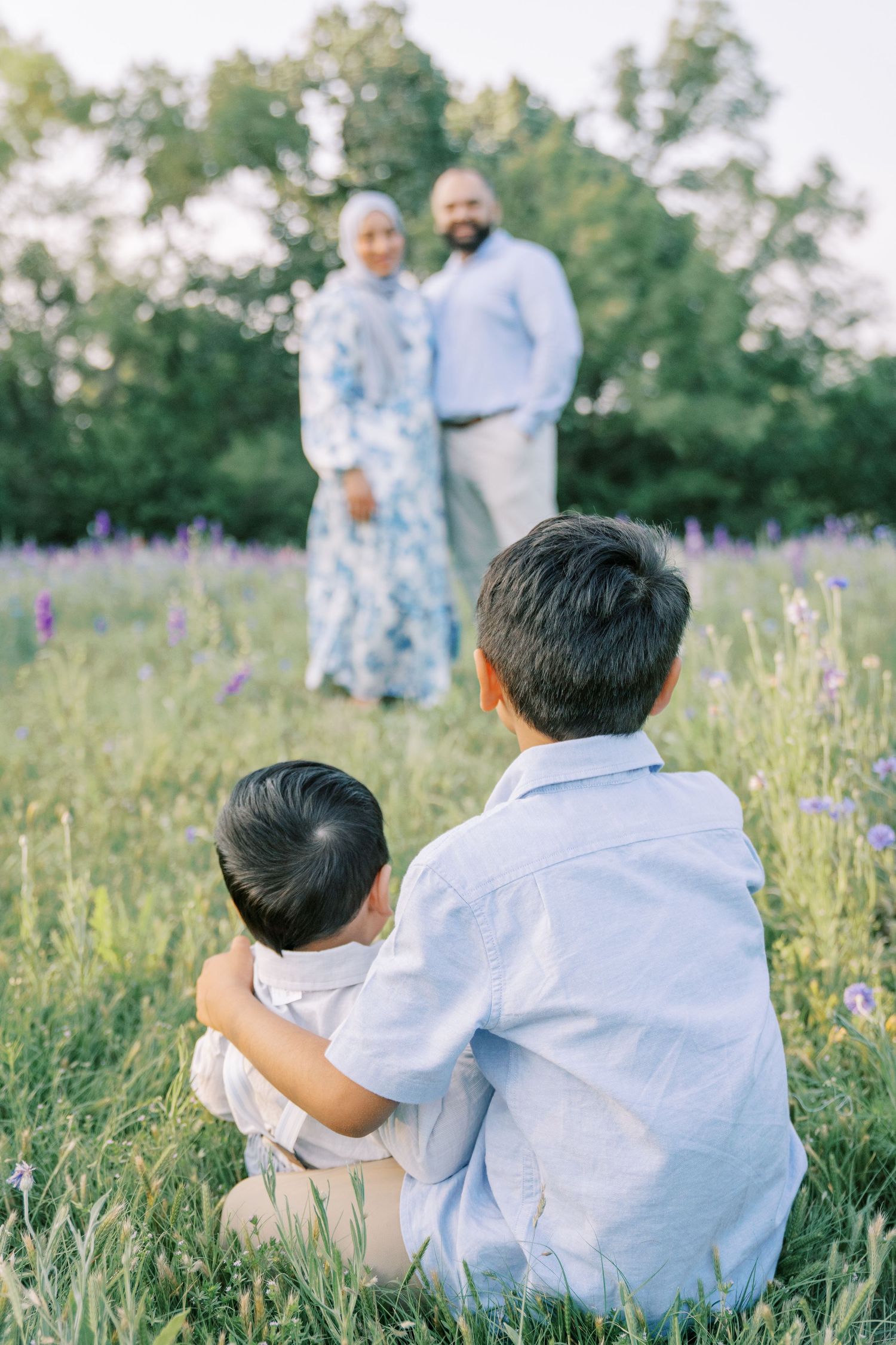 Two boys sit in a wildflower meadow facing their parents standing together among purple blooms at golden hour.
