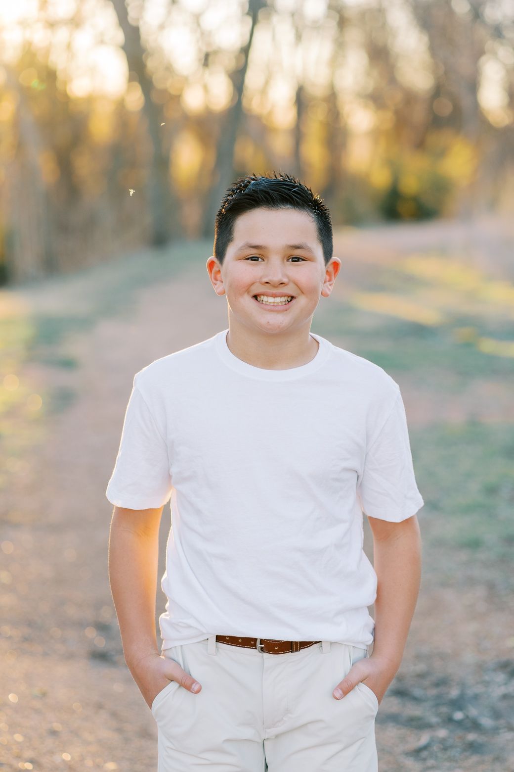 A young person in white clothing poses outdoors during golden hour in a natural setting.