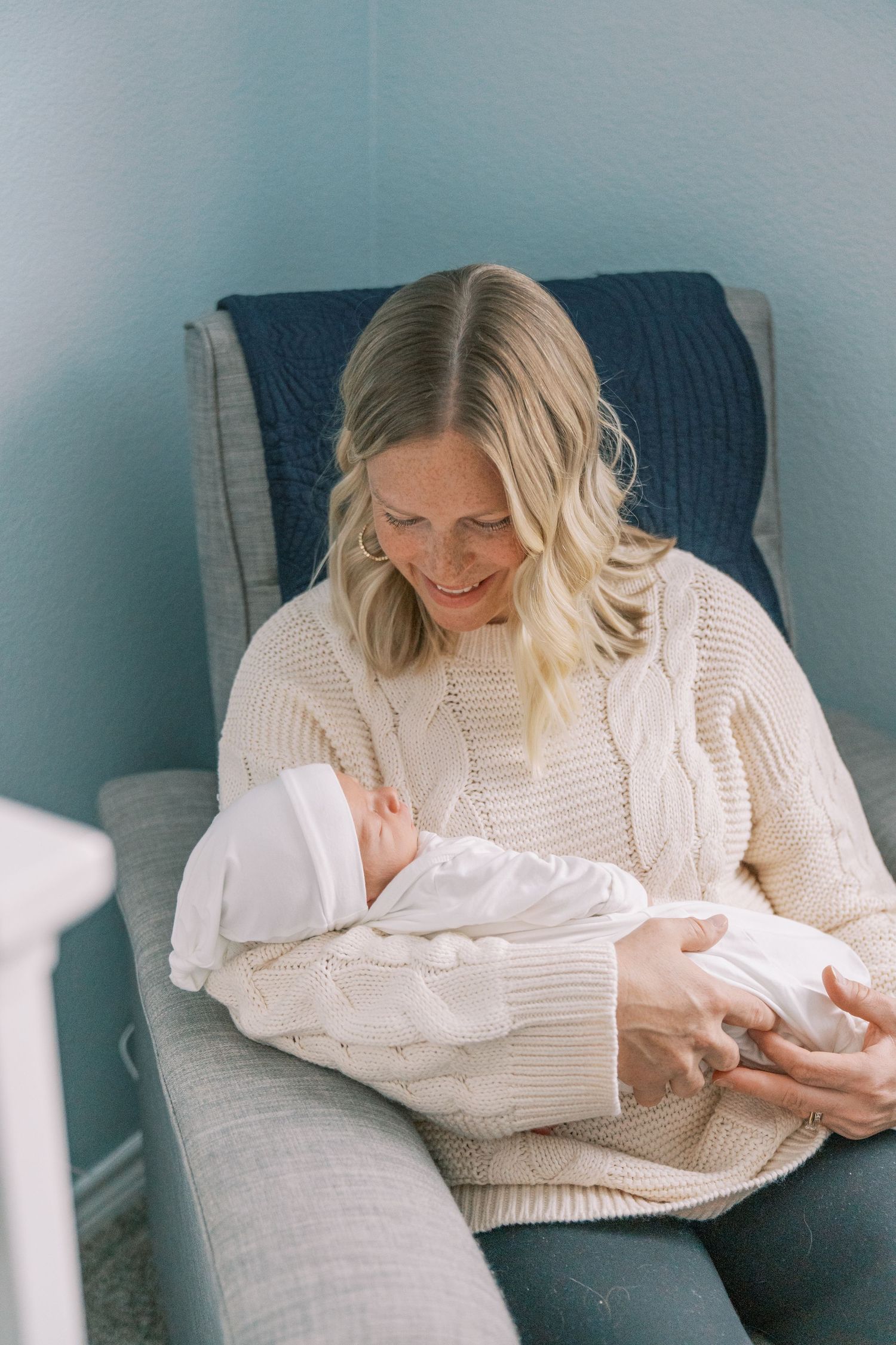 Parent in cream sweater tenderly holds sleeping newborn wrapped in white.