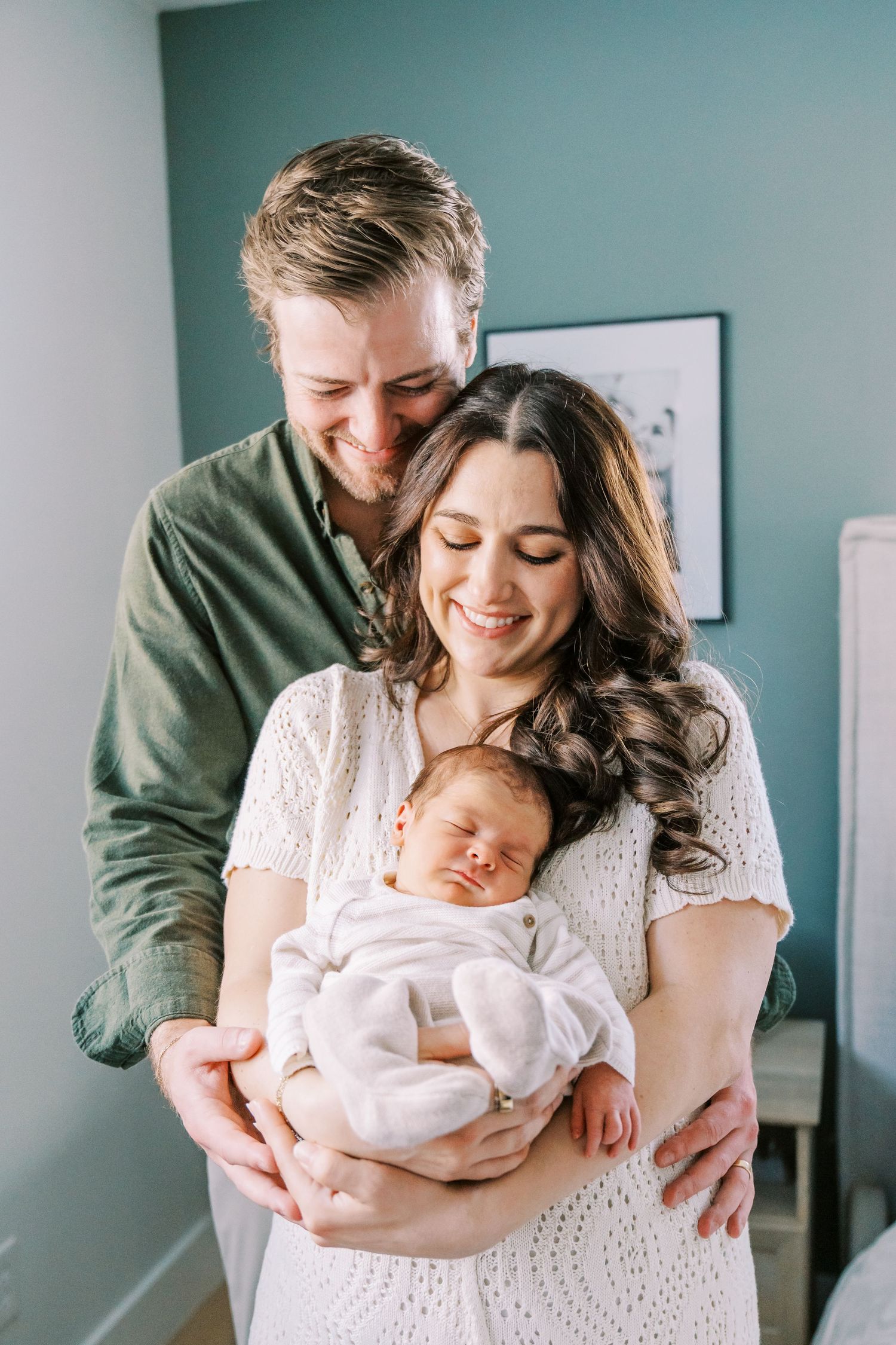 Parents in green and white outfits share tender moments with their newborn baby in a bright room.