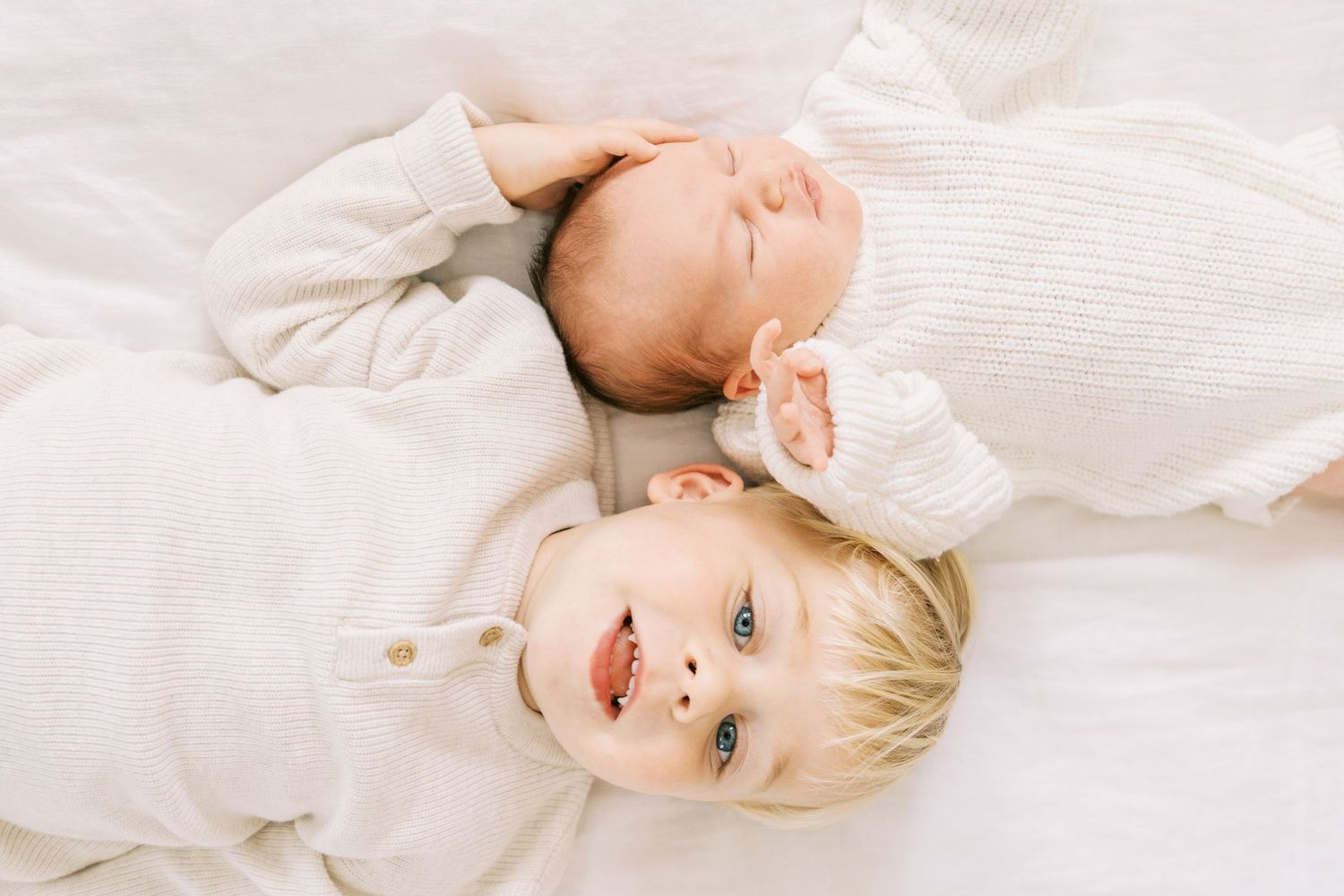 A baby and toddler lying together on white bedding in natural light.
