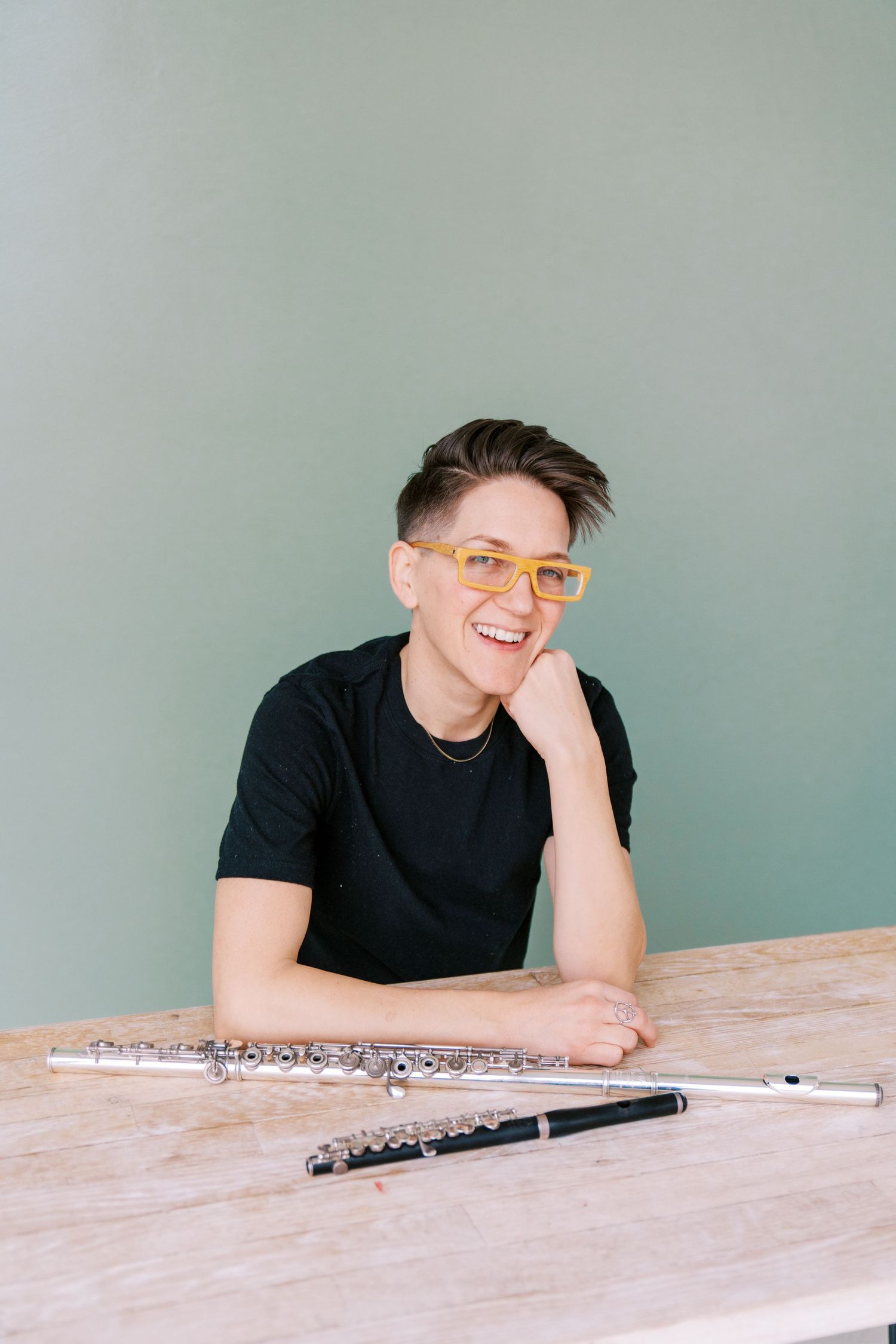 Musician in black shirt smiles while resting at wooden table with flute against mint green background.