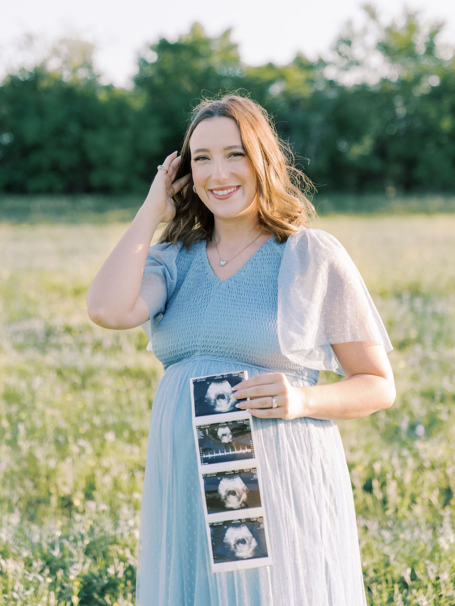 A woman in a light blue dress holds ultrasound photos while standing in a sunny wildflower field.