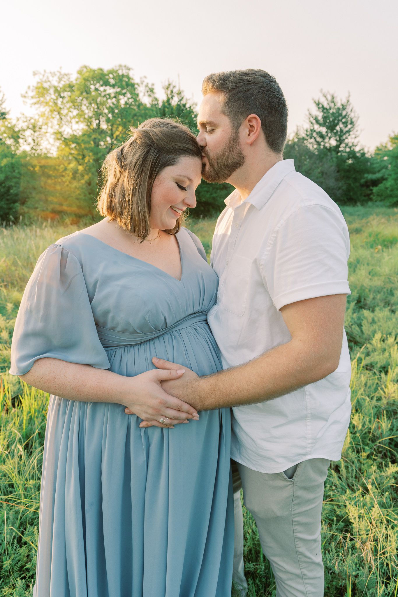 Tender moment between expecting couple in natural outdoor setting at sunset.