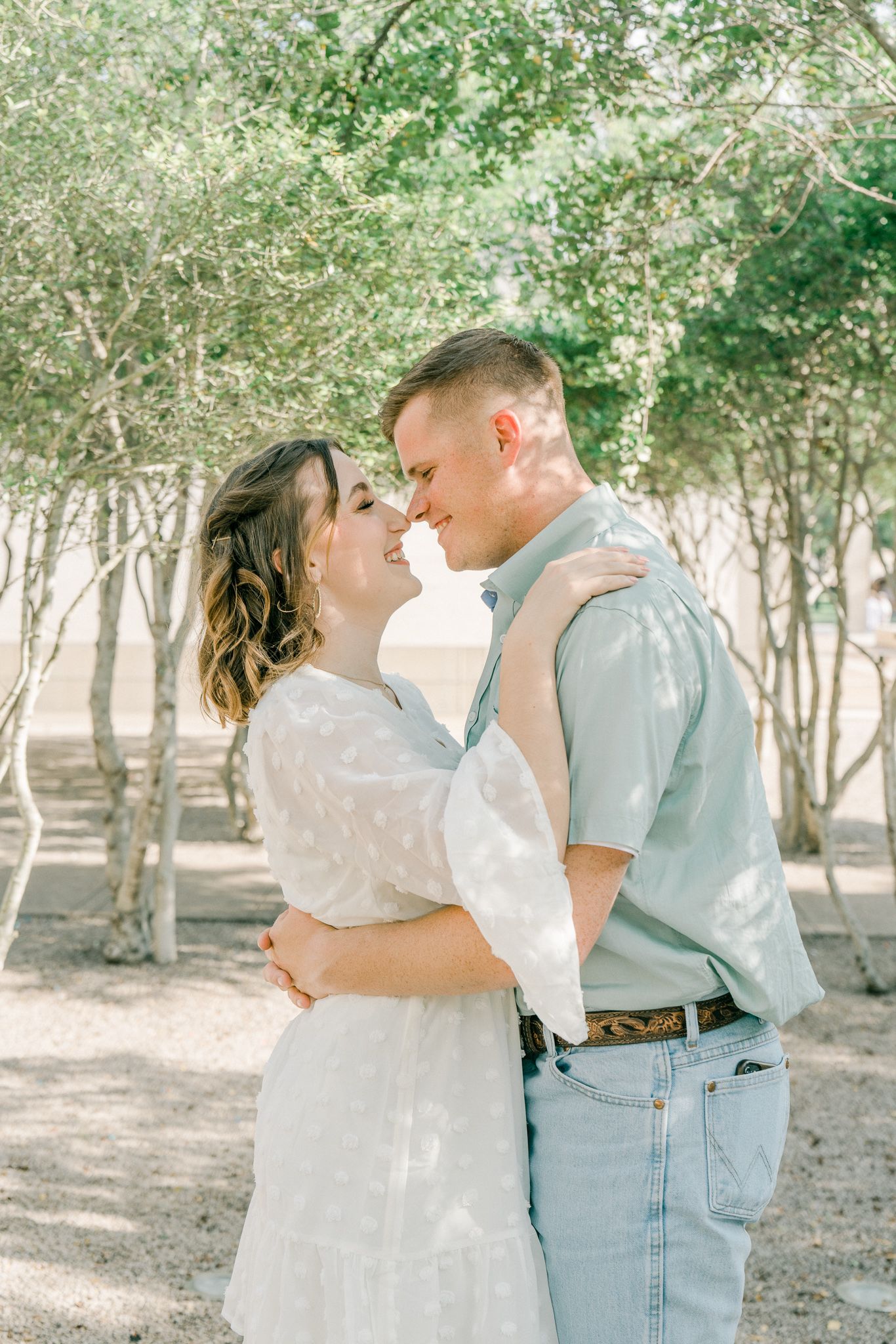 A romantic couple shares tender moments in a sunlit orchard wearing white and light blue outfits.