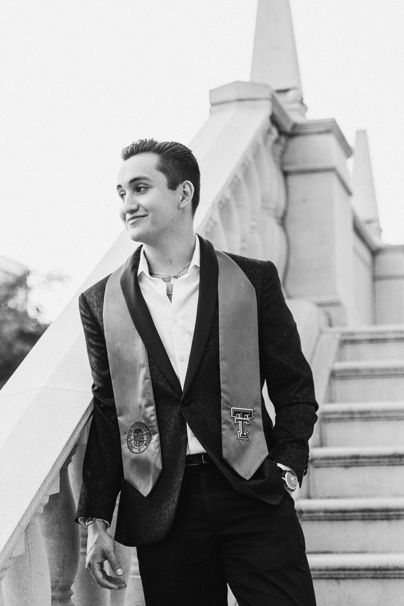 A graduate in black cap and gown with silver stole poses on stone steps outdoors.