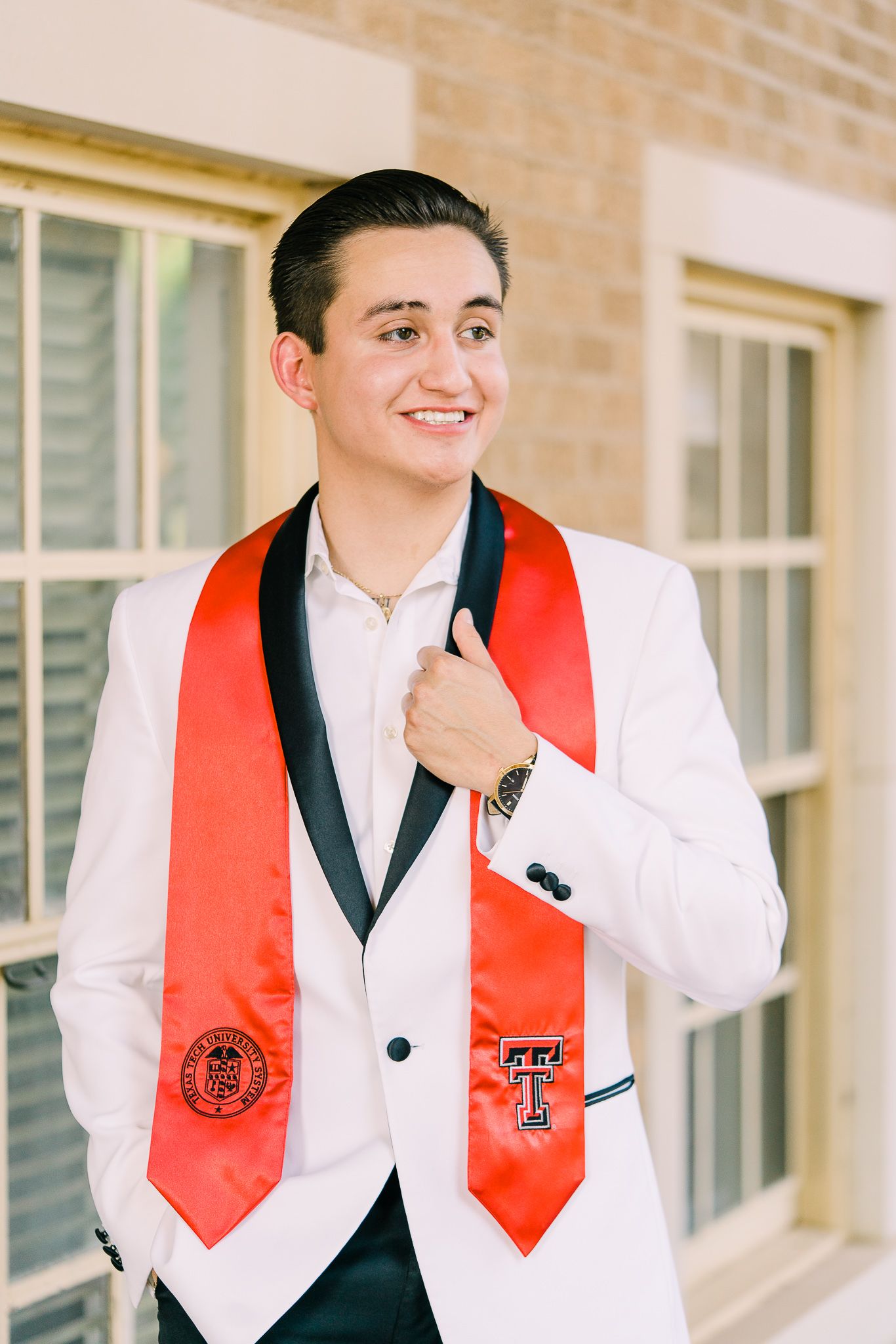 Graduate in white suit with red graduation stole poses in front of brick building.