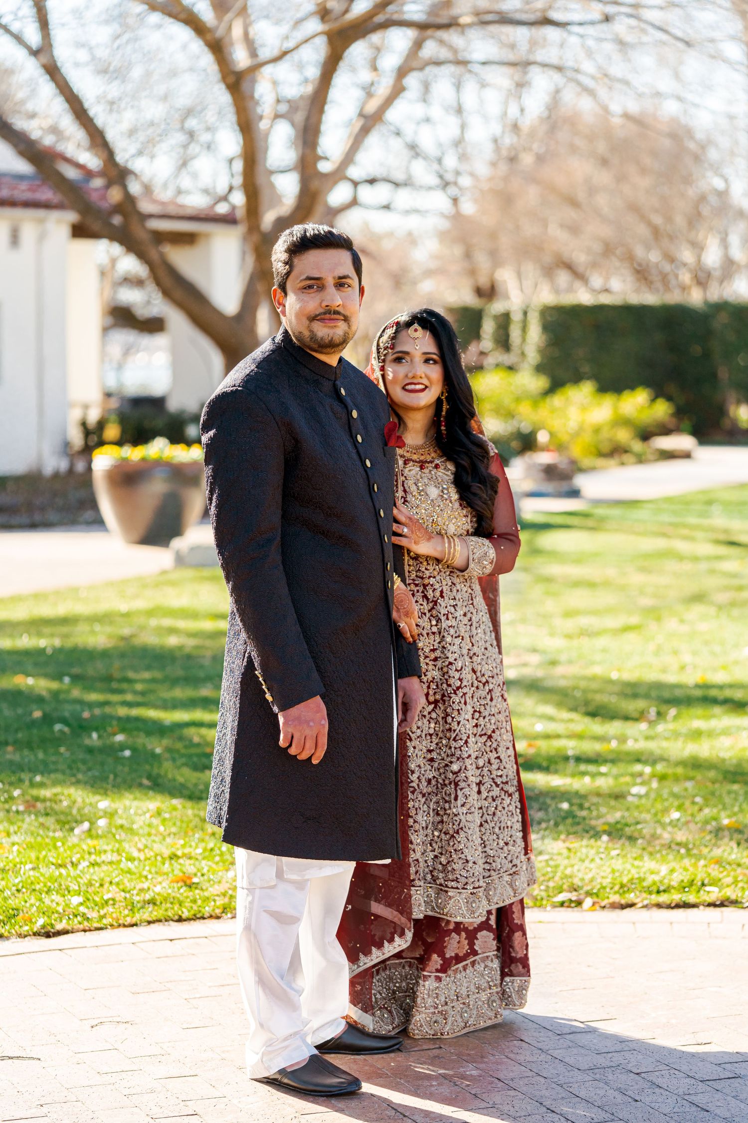 A couple in traditional South Asian attire poses outdoors with columns and trees in the background.